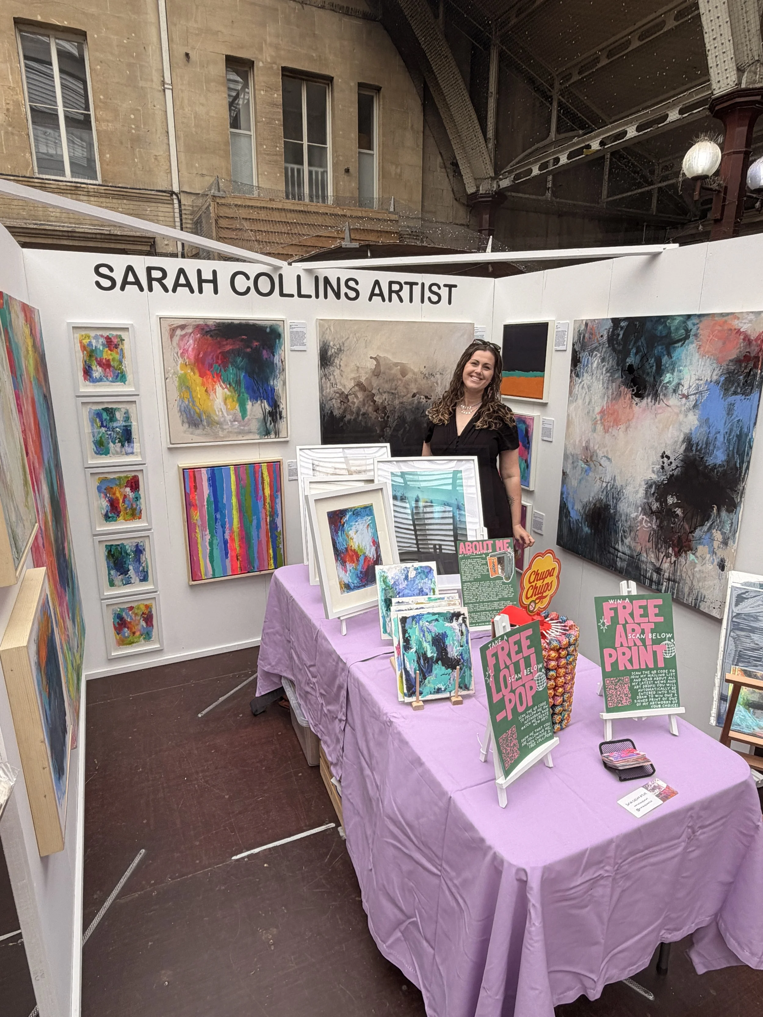 Art booth with paintings by Sarah Collins at an outdoor event. The artist, a woman with curly hair and black clothing, is smiling behind a table covered with pink cloth. The booth features colorful abstract paintings and promotional signs offering free prints.