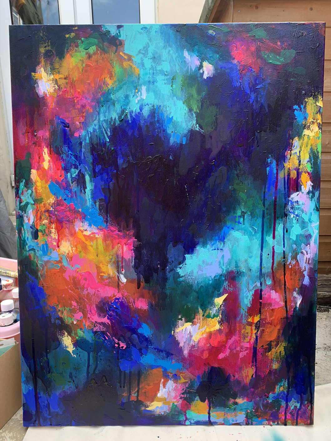 Abstract colorful painting with vibrant splashes of pink, orange, yellow, green, blue, purple, and black. Contains drips and textured areas on canvas.