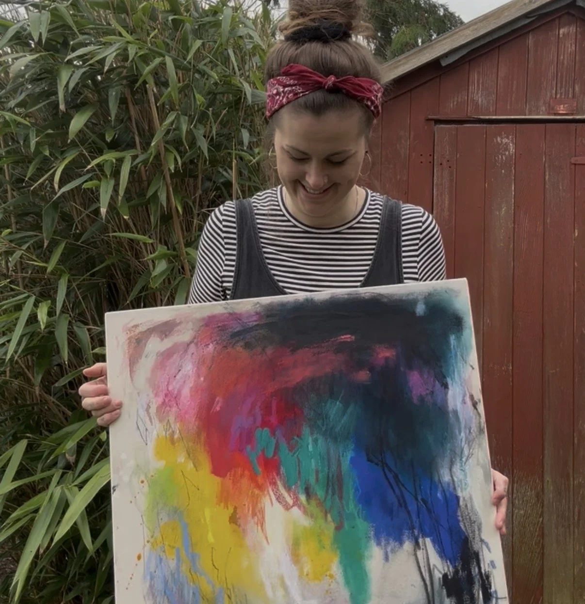 A woman with a red bandana in her hair, smiling while holding an abstract painting with vibrant colors, outdoors near green plants and a red wooden structure.