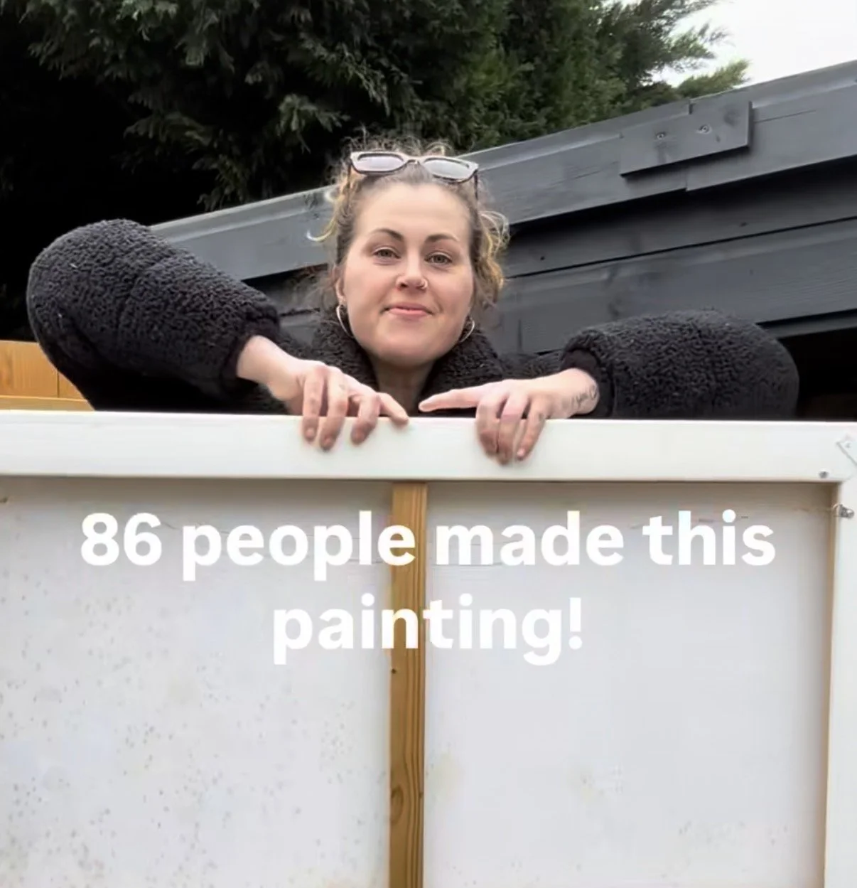 A woman with glasses on her head and earrings leaning on a white-framed painting outdoors, with trees in the background, and text overlay stating "86 people made this painting."