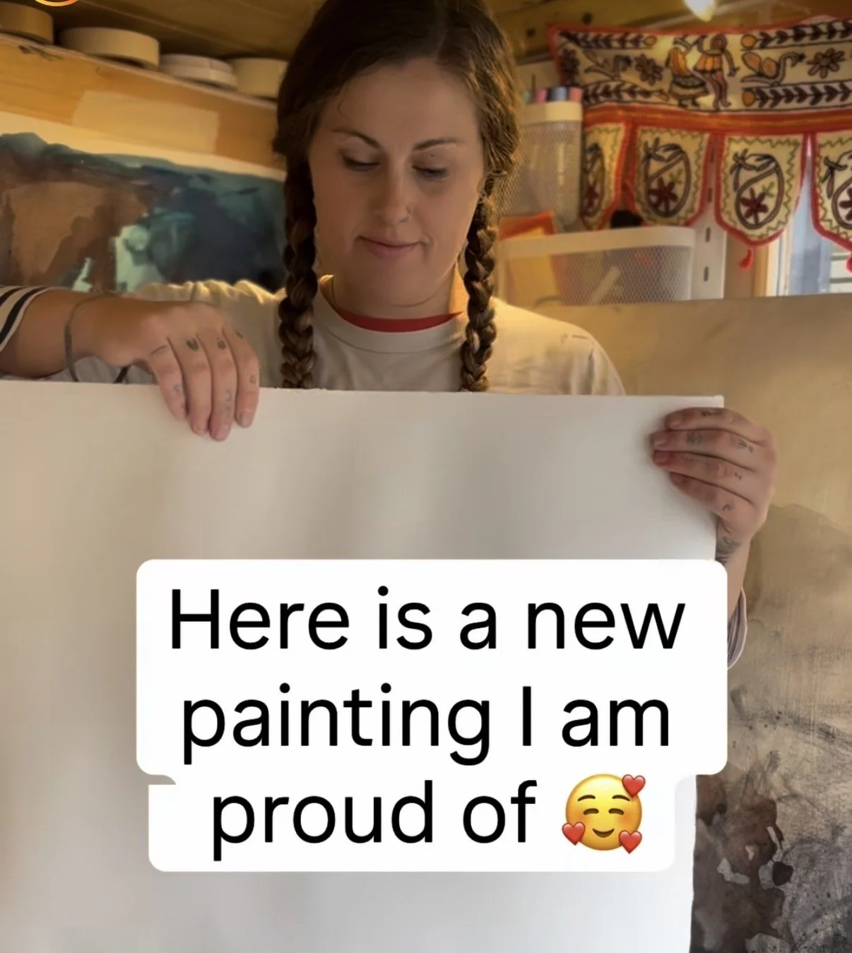 A woman with braided hair holding a blank canvas or paper, smiling slightly. The background shows colorful decorative items and containers. There is overlay text that says, 'Here is a new painting I am proud of 😍'.
