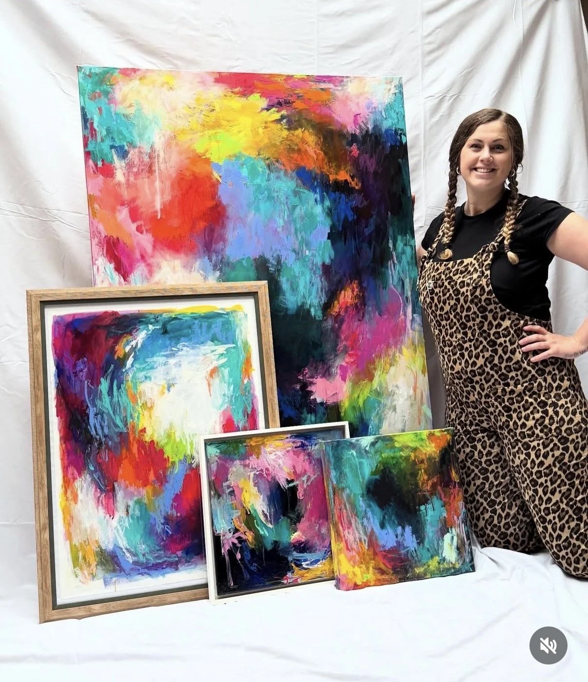 A woman with braided hair, wearing a leopard print apron over a black shirt, stands smiling next to a display of colorful abstract paintings. The paintings feature vibrant, bold splashes of color on various-sized canvases, some framed, set against a white backdrop.