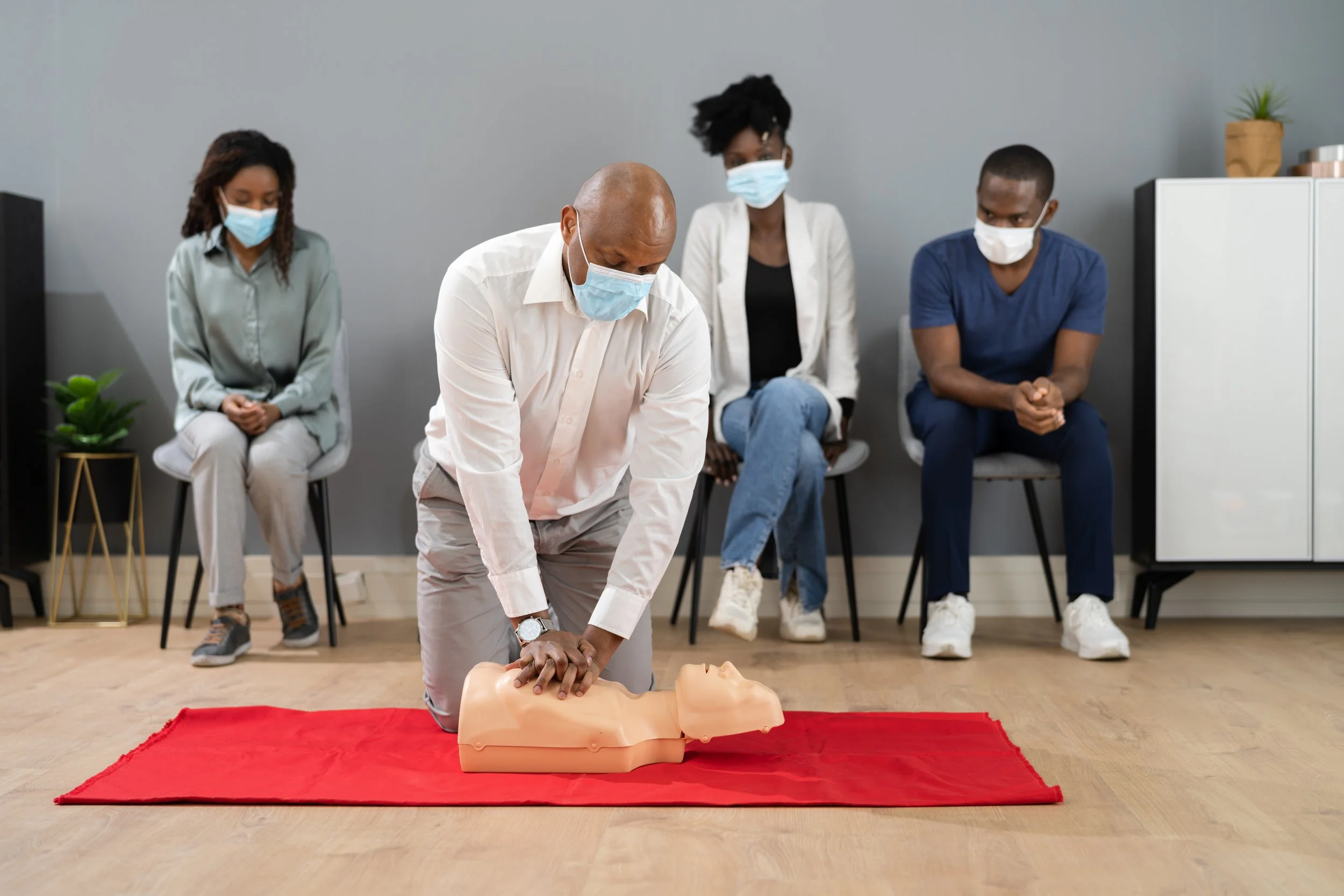 A man performs CPR on a training mannequin during a CPR training session, with three people in masks sitting on chairs in the background.