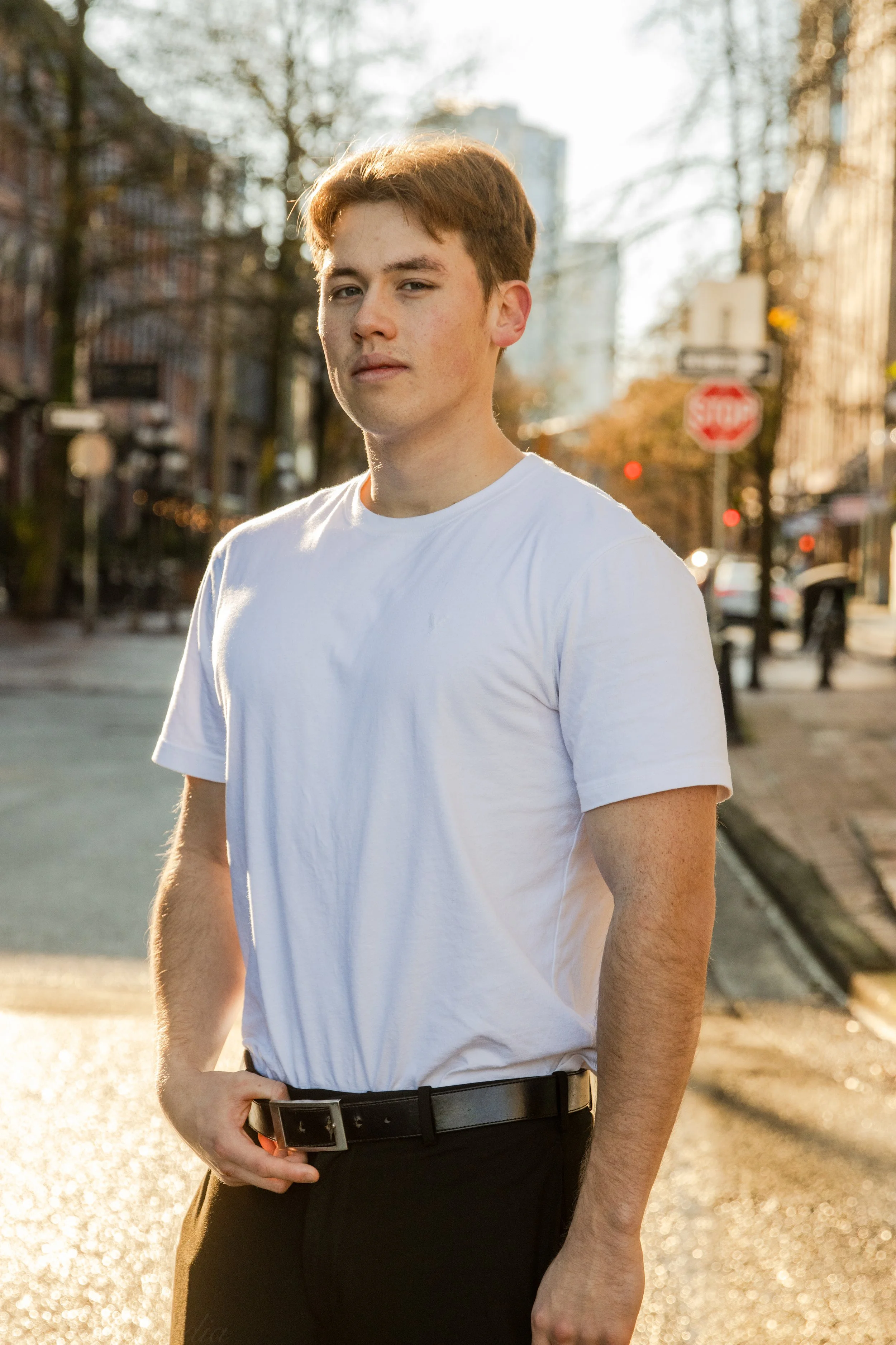 Young man in a white t-shirt and black belt standing on a city street during sunset.