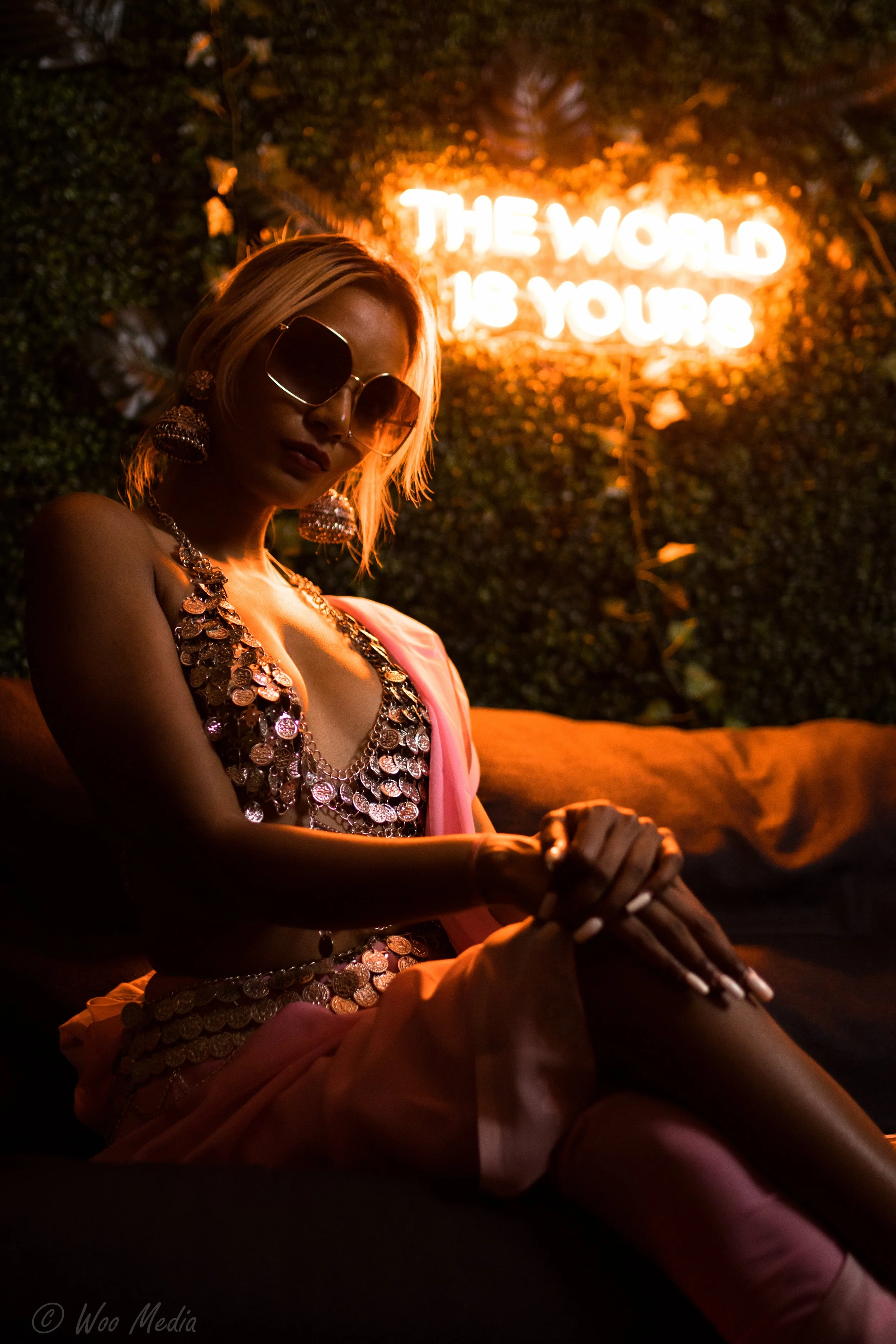 A woman with blonde hair wearing sunglasses and large earrings, sitting indoors with a dark background and an orange neon sign that reads 'The world is yours' behind her.