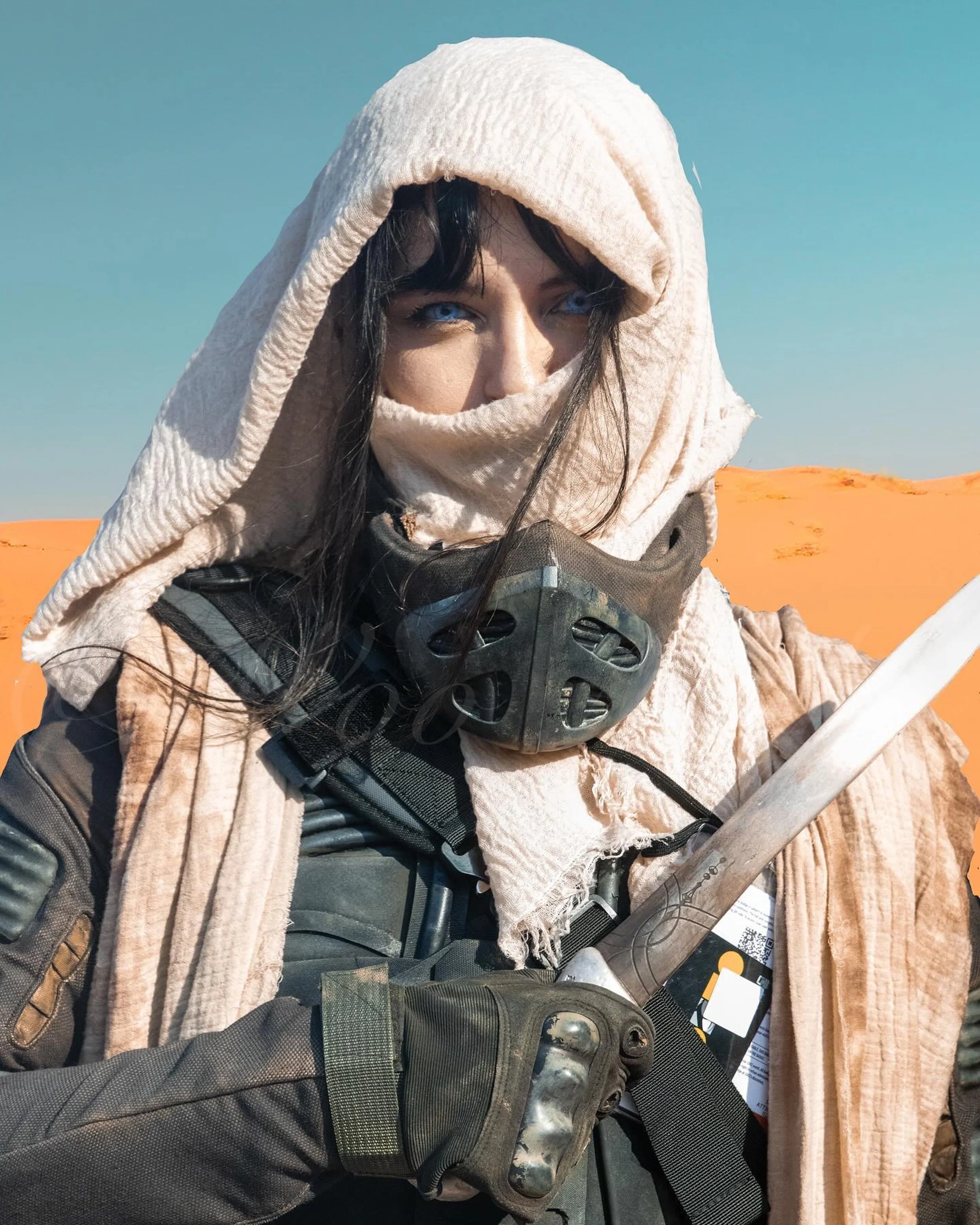 A person with blue eyes, wearing a beige headscarf and a black mask around their neck, dressed in outdoor tactical gear, holding a metal object, with a desert landscape in the background under a clear sky.