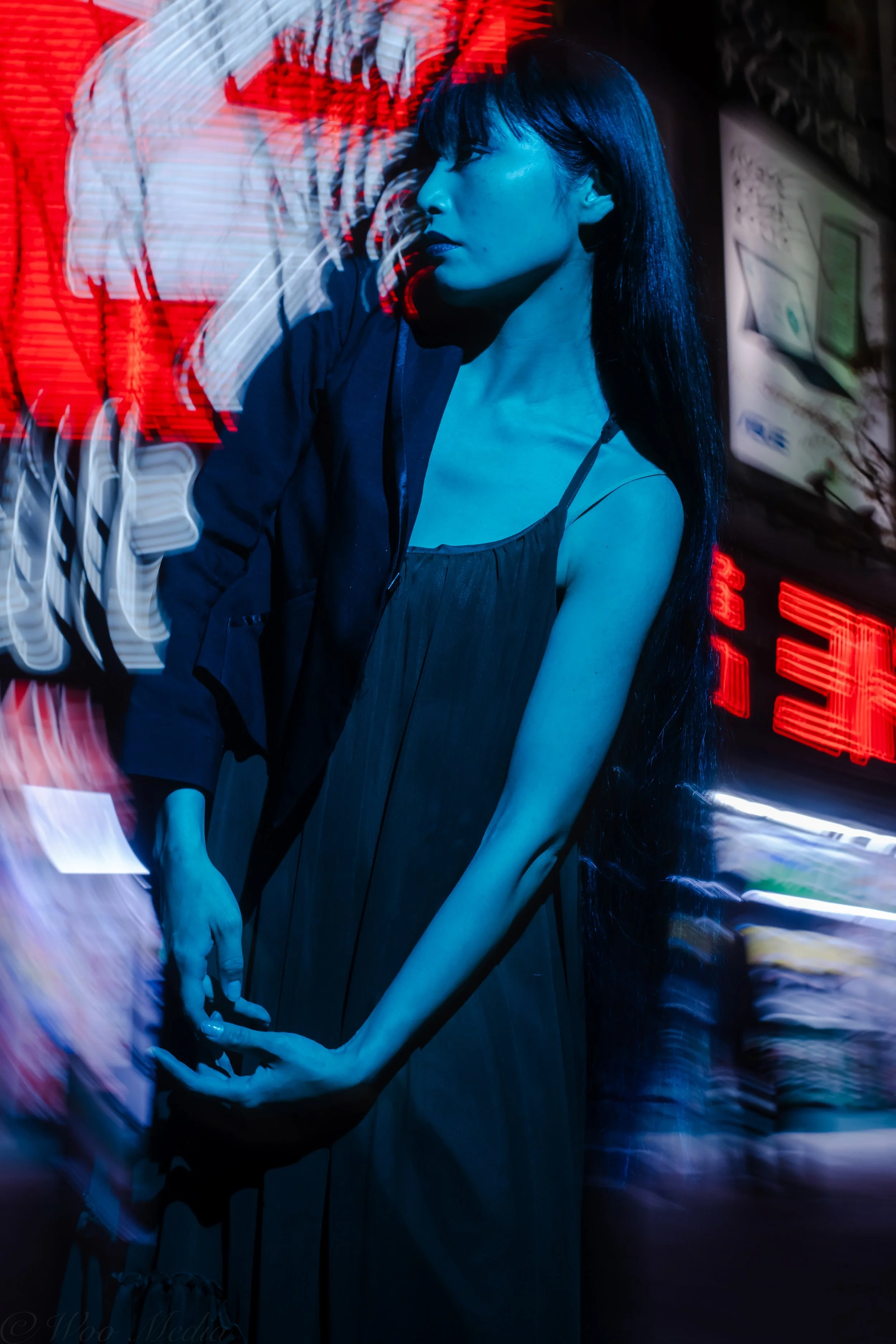 A woman with long black hair standing in front of a neon-lit sign, illuminated in blue and red light, with motion blur effects.