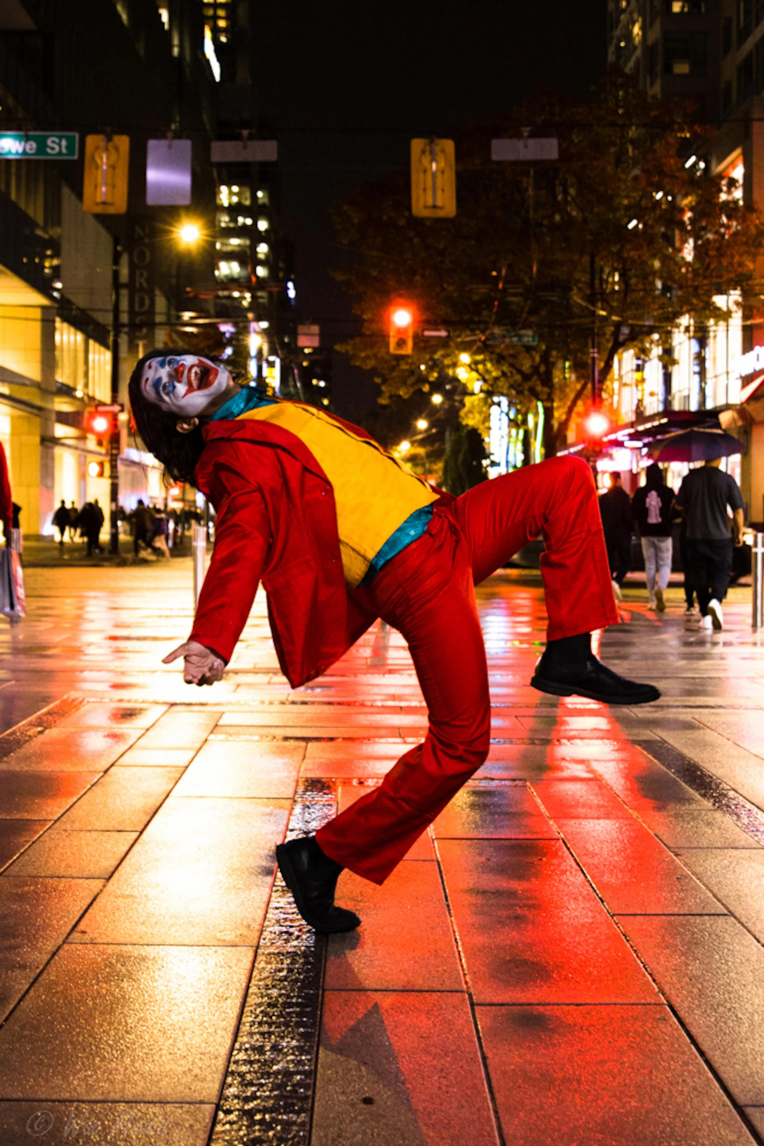 A person dressed as a clown with face paint, wearing a red jacket and pants, is dancing on a wet city street at night, illuminated by streetlights and signs, with pedestrians walking in the background.