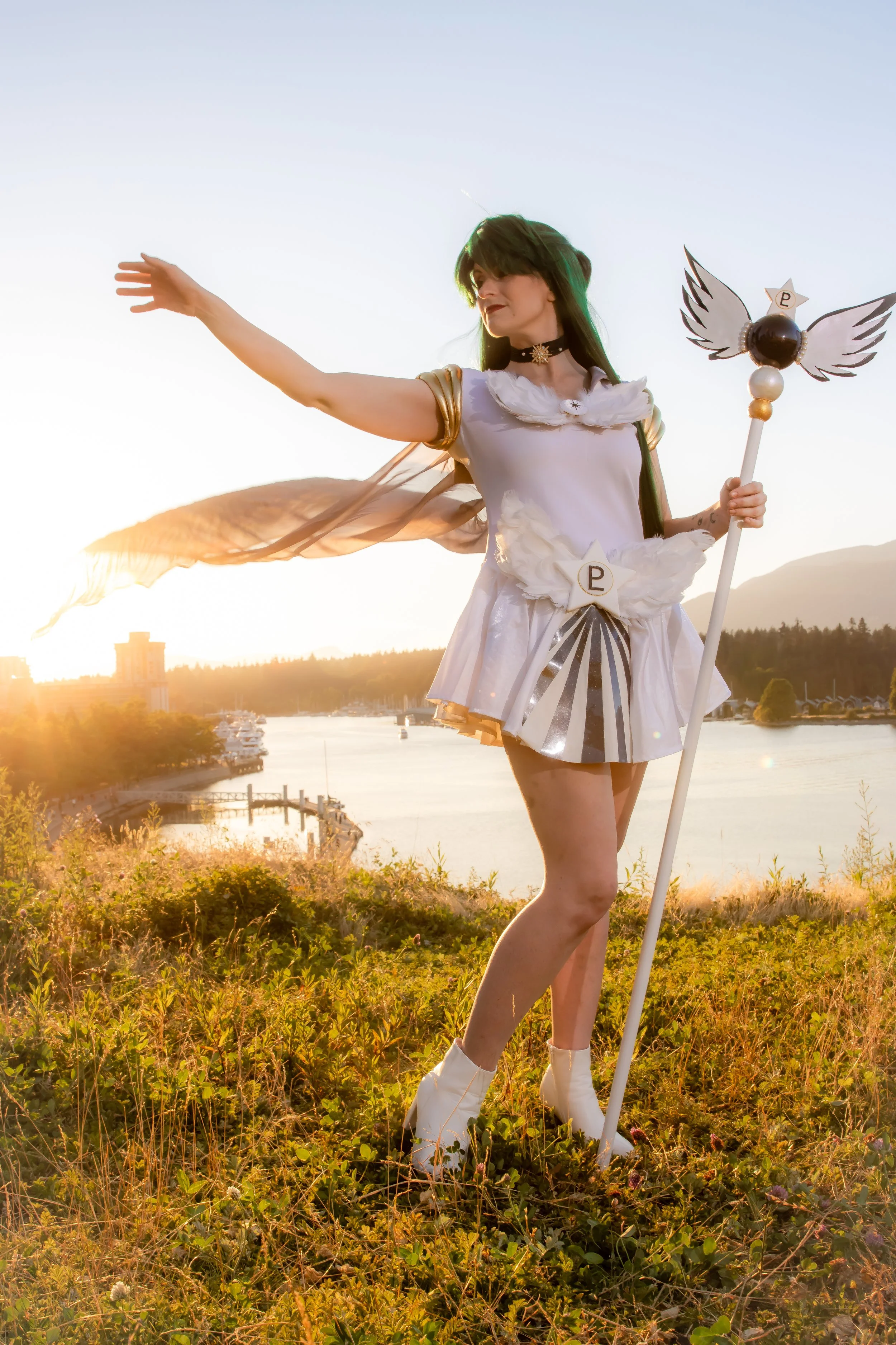 A person dressed as Sailor Moon standing outdoors during sunset, holding a staff with winged ornaments, with a scenic water and mountain background.