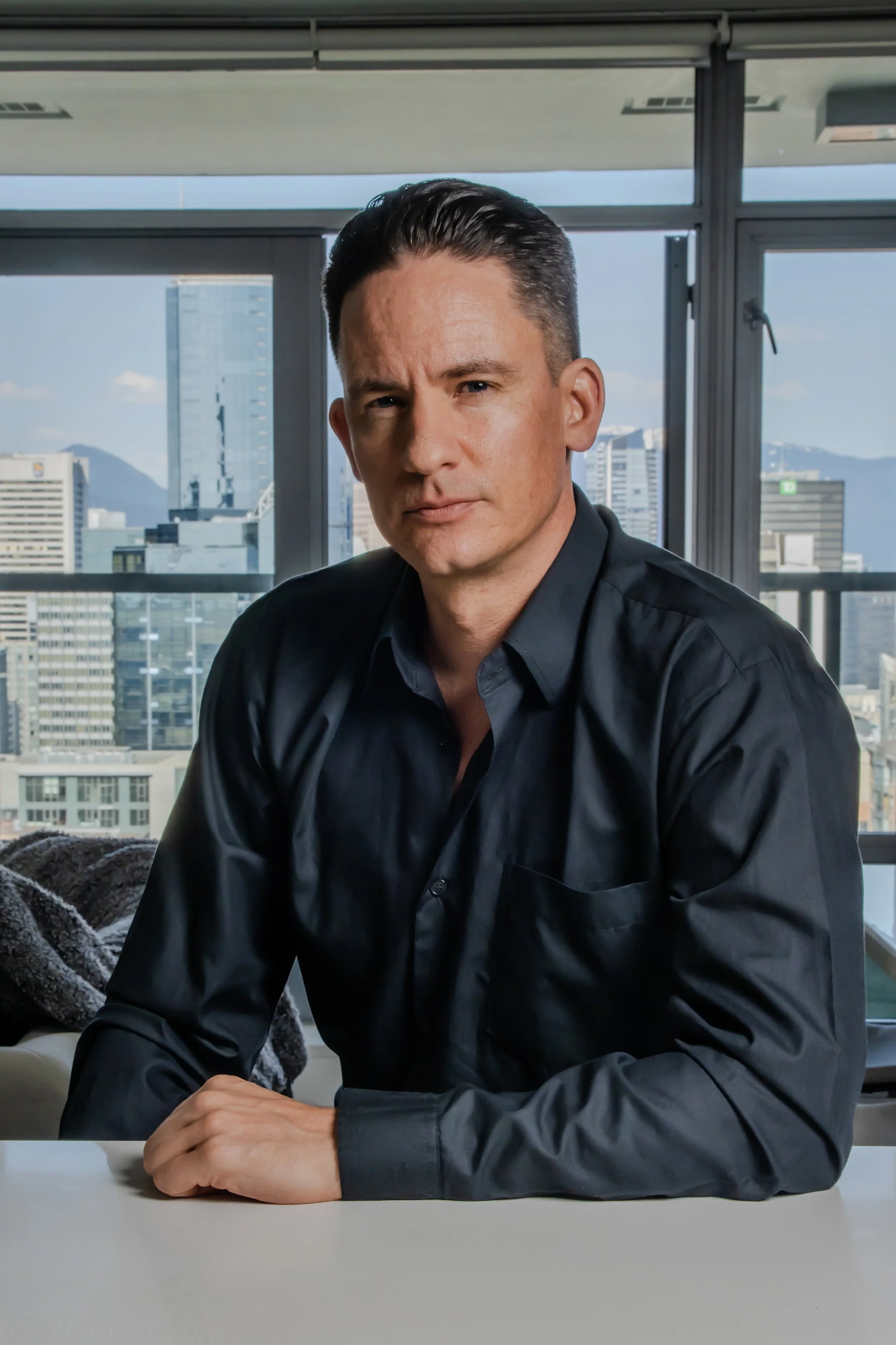 A man with slicked-back dark hair wearing a black dress shirt seated at a white table, with a cityscape view of tall buildings and mountains in the background through large windows.
