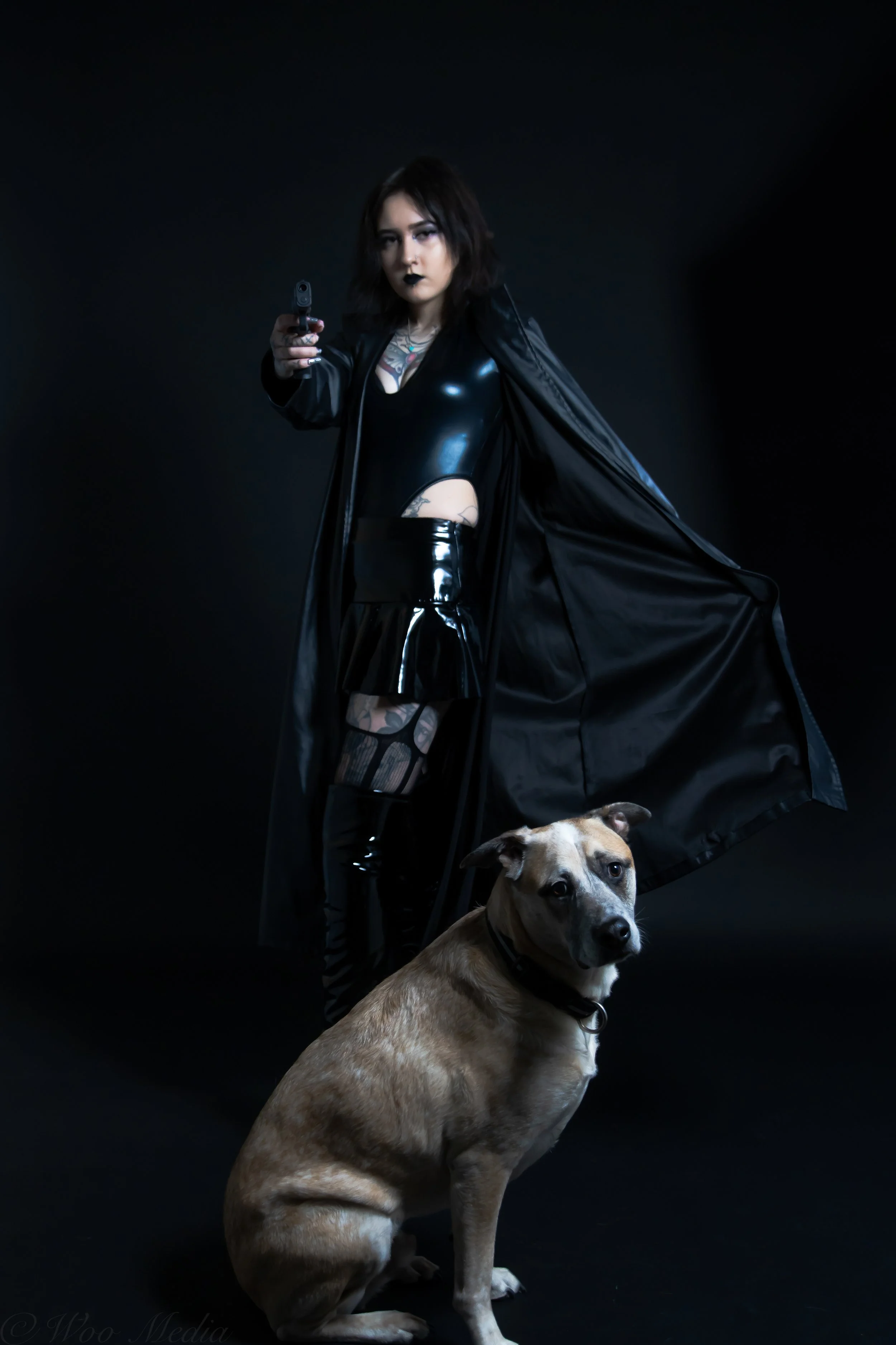 A woman dressed in black leather and vinyl with a gun pointed forward, standing against a black background, accompanied by a seated dog.