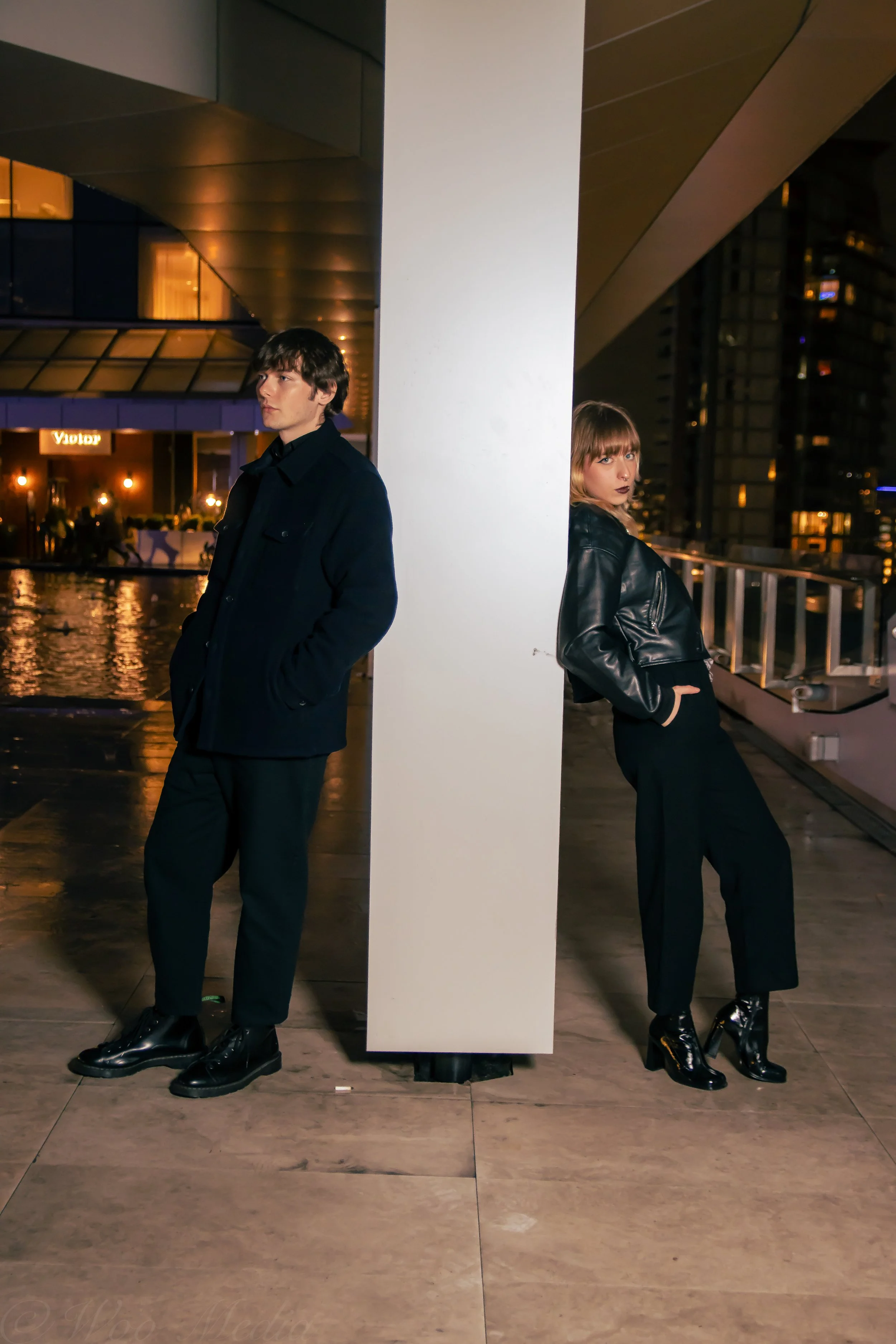 A man and a woman are standing on opposite sides of a white pillar on a rooftop at night. They are both wearing dark clothing and appear to be leaning against the pillar, looking in different directions. The background shows a cityscape with lit buildings and a reflecting pool.