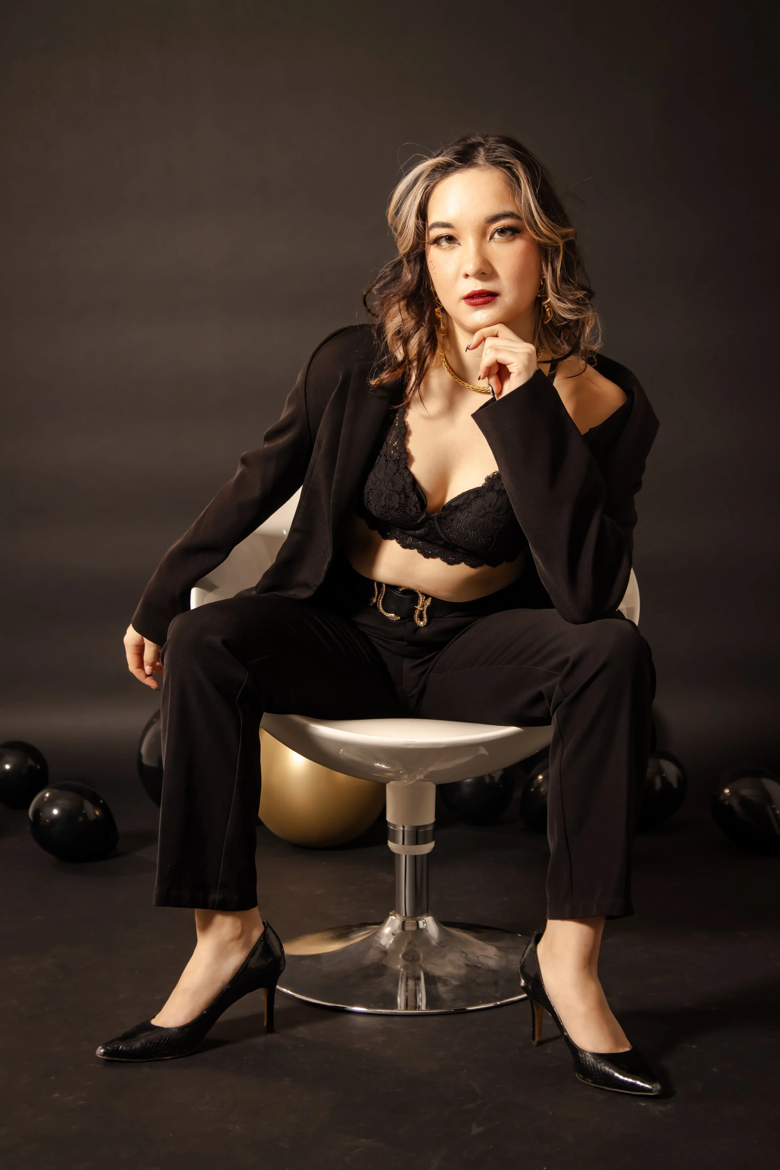 A woman with wavy hair, wearing a black lace bra, black blazer, black pants, and black heels, sitting on a white chair with one leg crossed over the other against a dark background with black balloons and a golden balloon