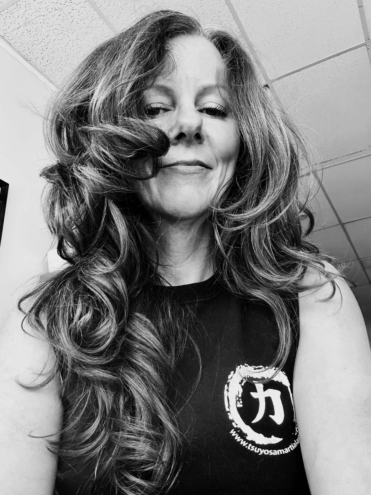 A woman with long, curly hair takes a selfie in a room with a tiled ceiling, wearing a sleeveless black top with a logo and website on it.