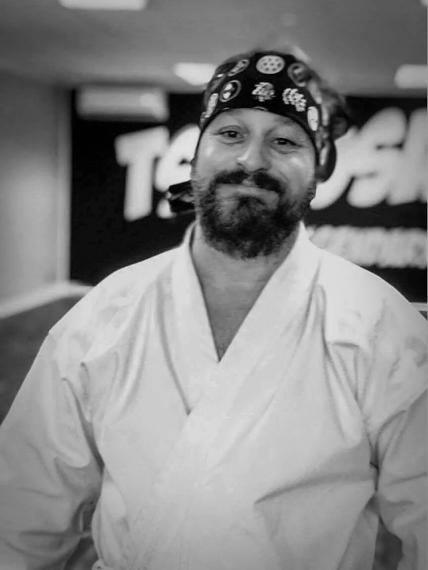 A man in a martial arts uniform and bandana smiling at the camera, with a blurred background and a partial logo or sign. Sensei Steve Derai.