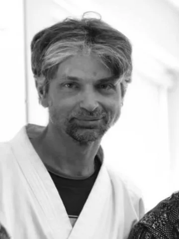 A man with medium-length hair and a beard, smiling, wearing a white shirt. Sensei at Tsuyosa Martial Arts.