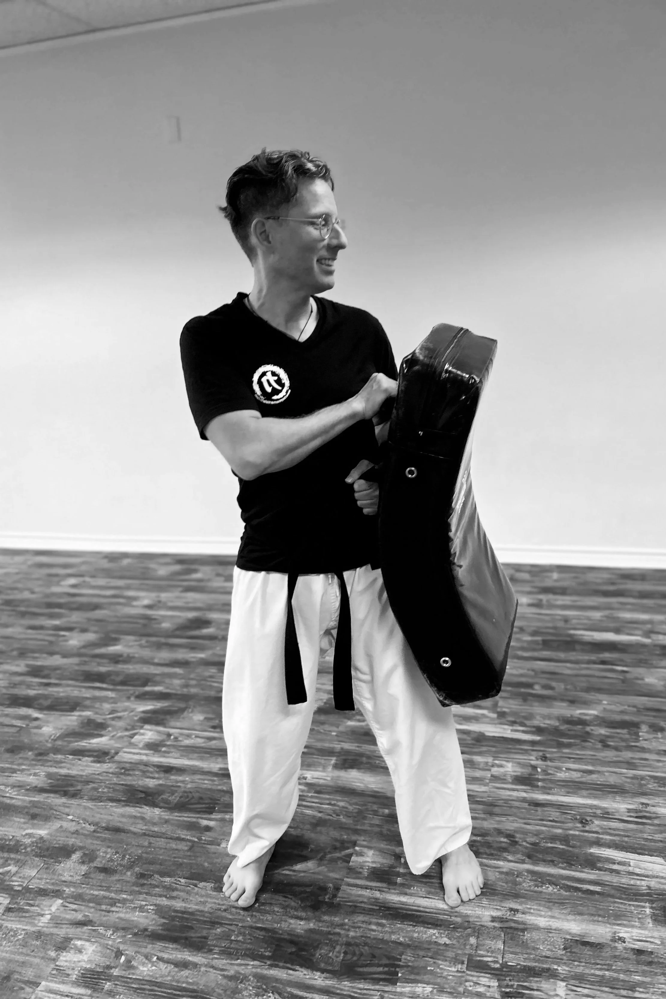 Person standing barefoot on wooden floor, smiling, holding a black shield, wearing a black t-shirt and white martial arts pants.