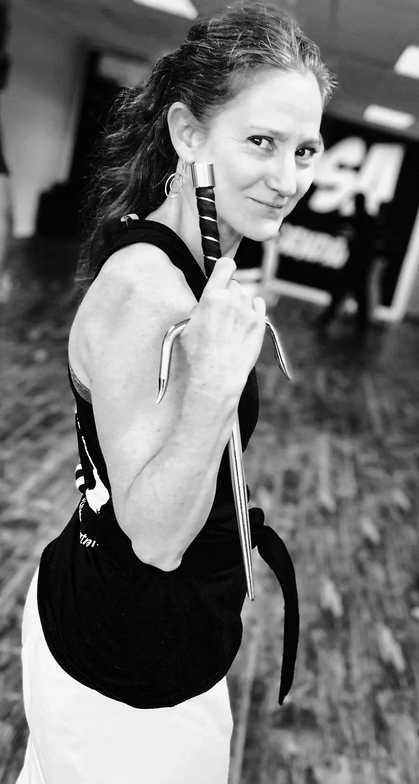 A woman holding a sai weapon in a martial arts studio, smiling at the camera, in black and white.