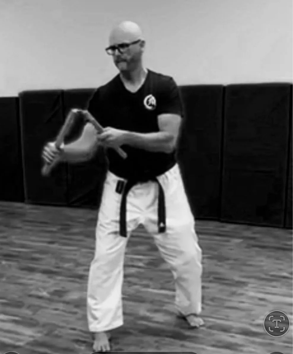 A man practicing martial arts in a dojo, wearing a martial arts gi with a black belt and holding a wooden training weapon. Sensei at Tsuyosa Martial Arts.