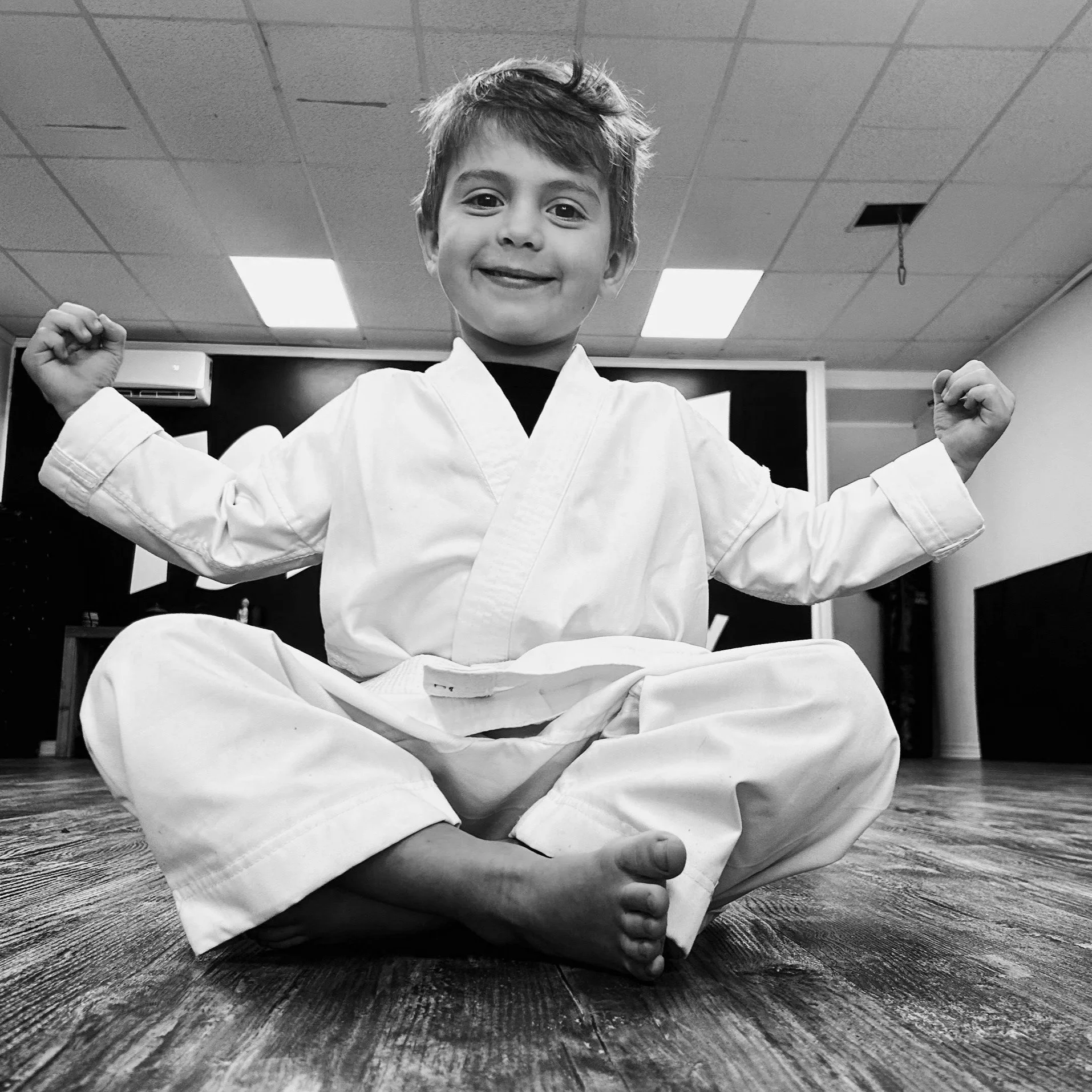 A young boy in a martial arts uniform seated in a cross-legged pose, smiling and flexing his arms in a dojo or gym setting.