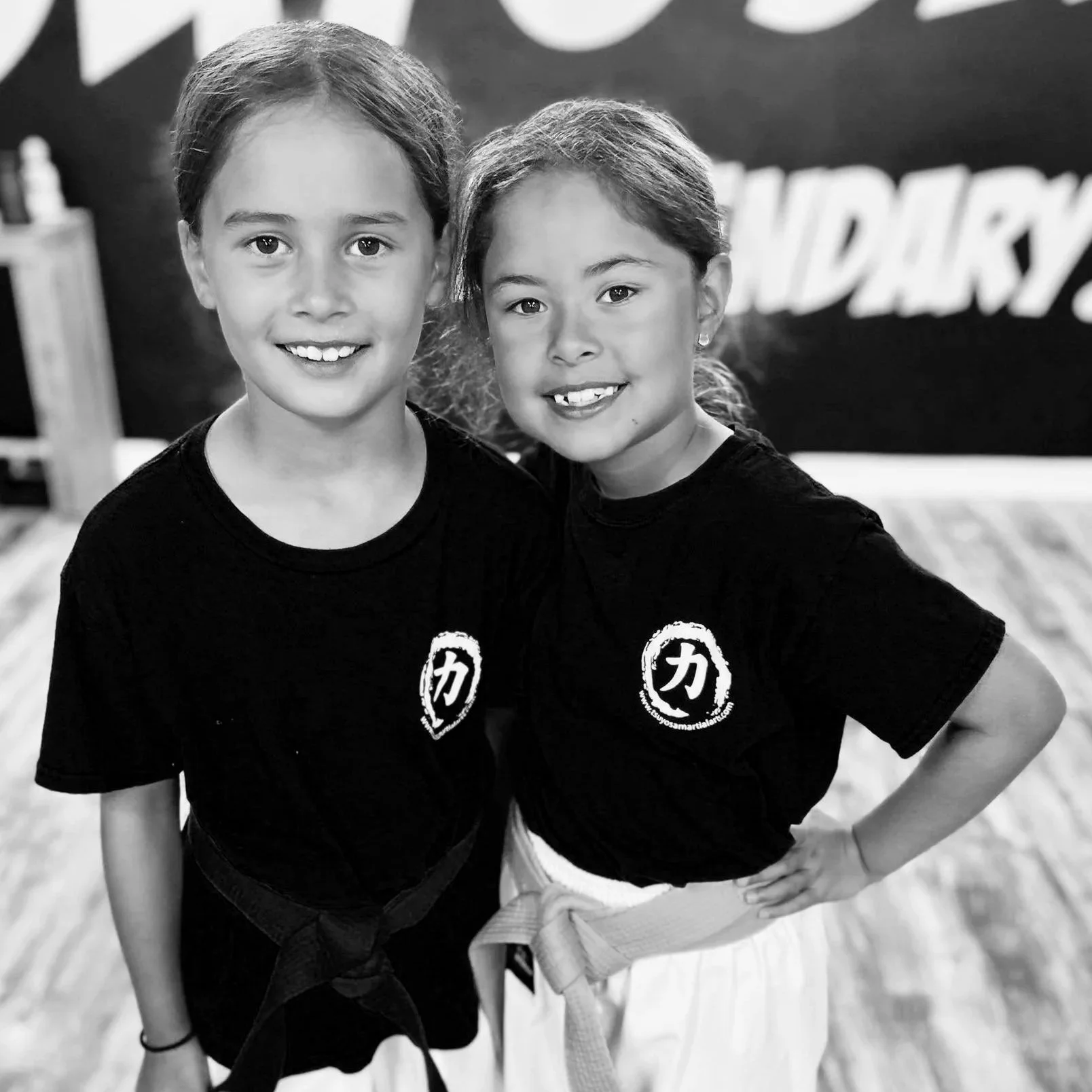 Two young girls wearing black t-shirts with martial arts logos, smiling, at a martial arts class or event.