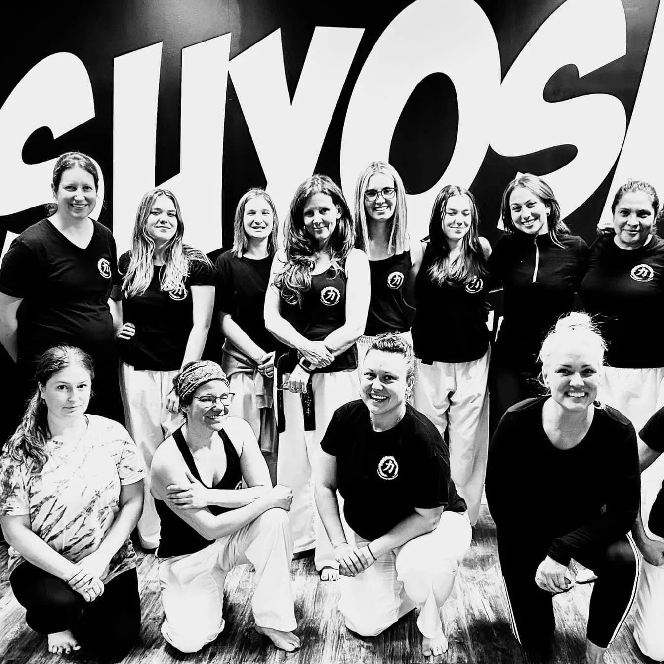 Group of women in martial arts uniforms and black T-shirts posing together inside dojo after a self defence workshop.