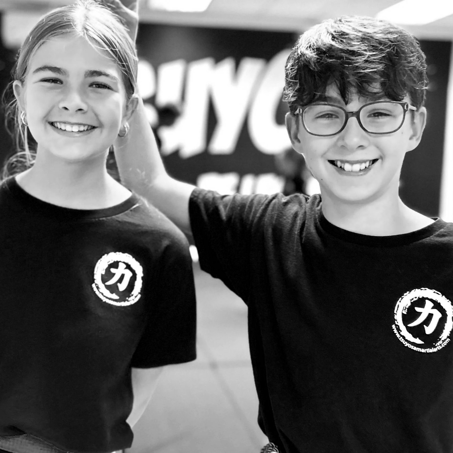 A smiling girl and boy standing side by side with the girl having her arm around the boy, both wearing black T-shirts with a logo on the chest. The girl has light hair and the boy has curly hair and wears glasses. Enjoying teen martial arts.