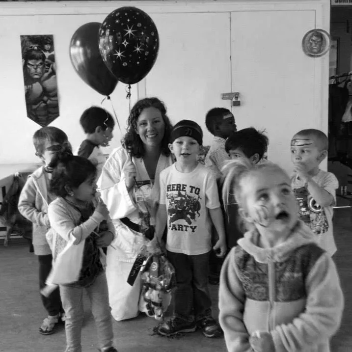A woman dressed as a karate expert celebrating with children at a party with balloons, some children are smiling and looking surprised.