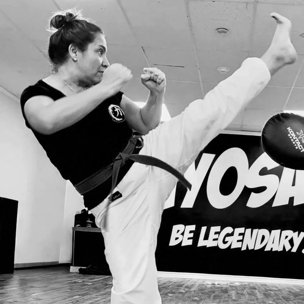 Woman practicing martial arts in a gym, performing a high kick, wearing a black shirt and white gi with a black belt, indoor training space with a sign that says 'BE LEGENDARY'.