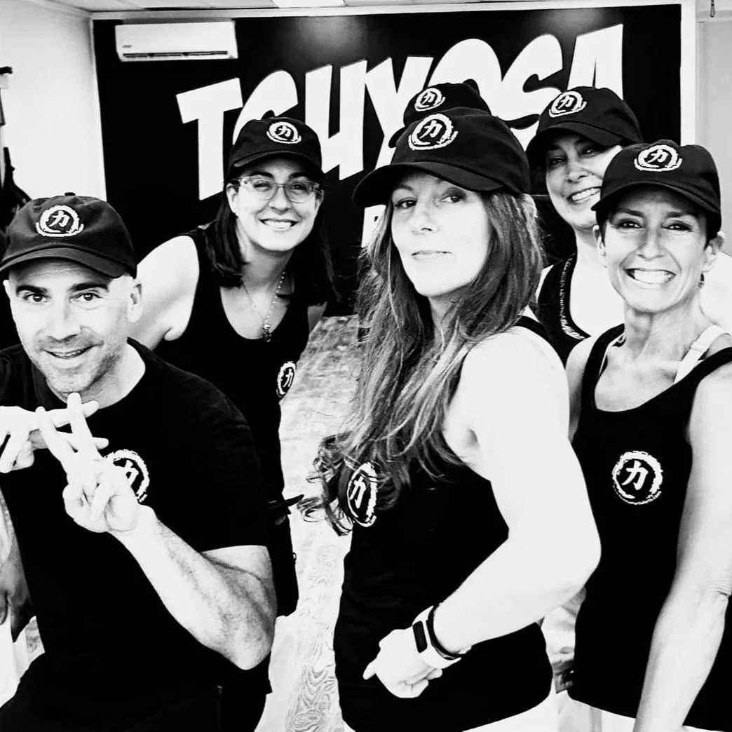 Group of six smiling people wearing matching black hats and shirts with a logo, standing in front of a sign that says 'TSUYOSA' in an indoor setting.