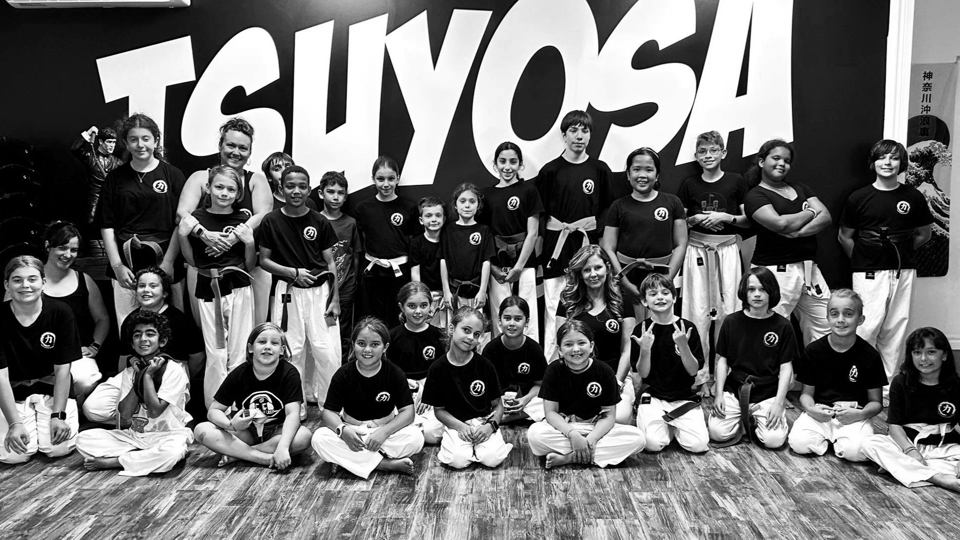 Group of children and two instructors in martial arts uniforms posing for a photo in front of a large 'TSUYOSA' sign. Some children are kneeling in the front, others standing in the back.