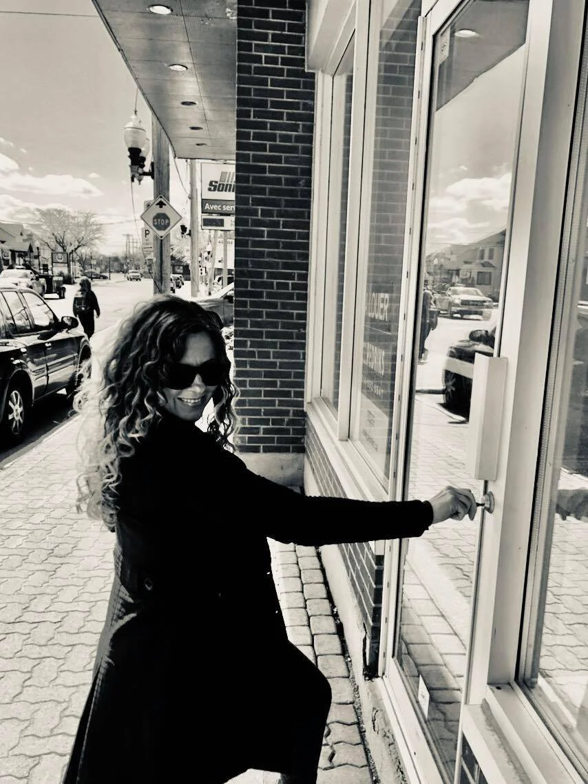 Woman with curly hair and sunglasses opening a glass door outside a storefront.