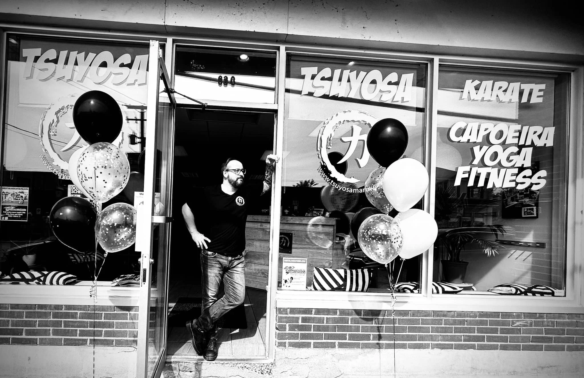 Man standing in front of a fitness center decorated with balloons, large windows displaying signs for Tsuyosa martial arts, karate, capoeira, yoga, and fitness in bold letters, with reflections visible in the glass.