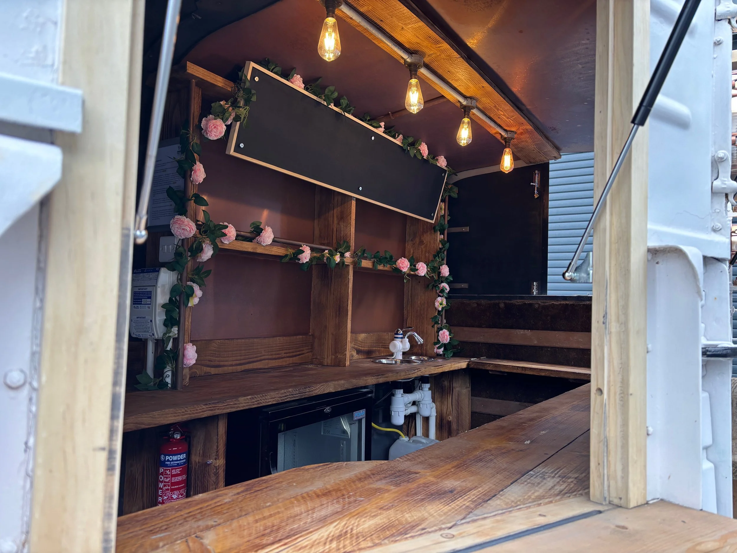 Interior of a small wooden food truck with shelves, a small sink, decorations of pink roses and a chalkboard, and warm light bulbs.