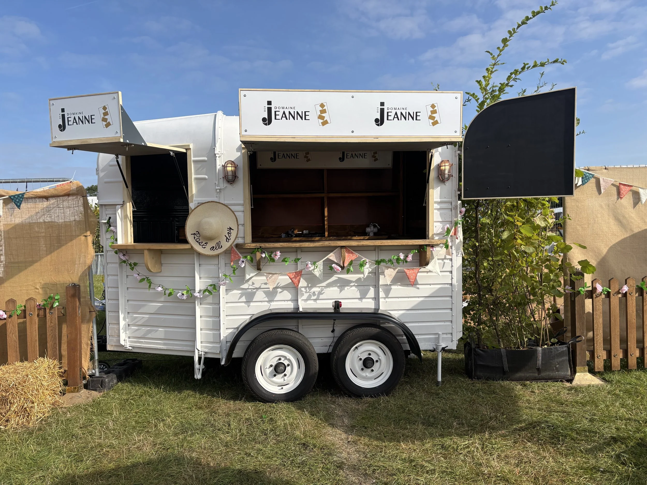 A small white food trailer with open service windows, decorated with bunting and green garland, branded with 'Domaine Jeanne', set outdoors with a fence, plants, and a blue sky.