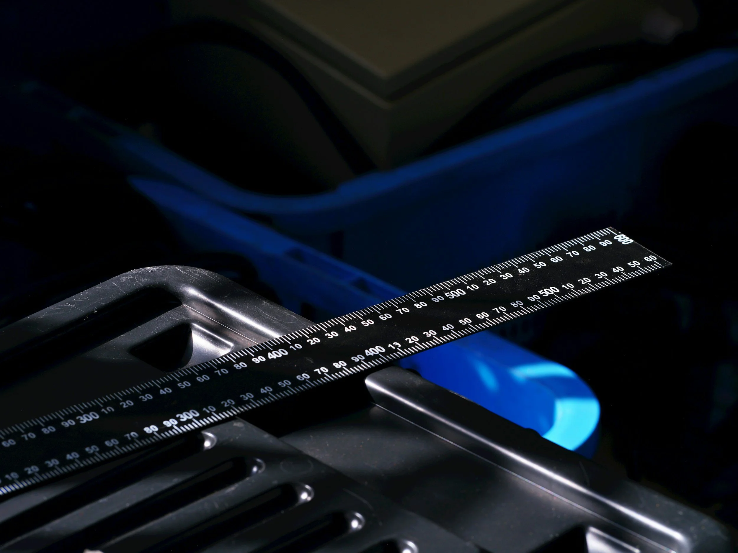 Close-up view of a black ruler measuring object on a metallic surface in a dark environment with faint blue lighting.