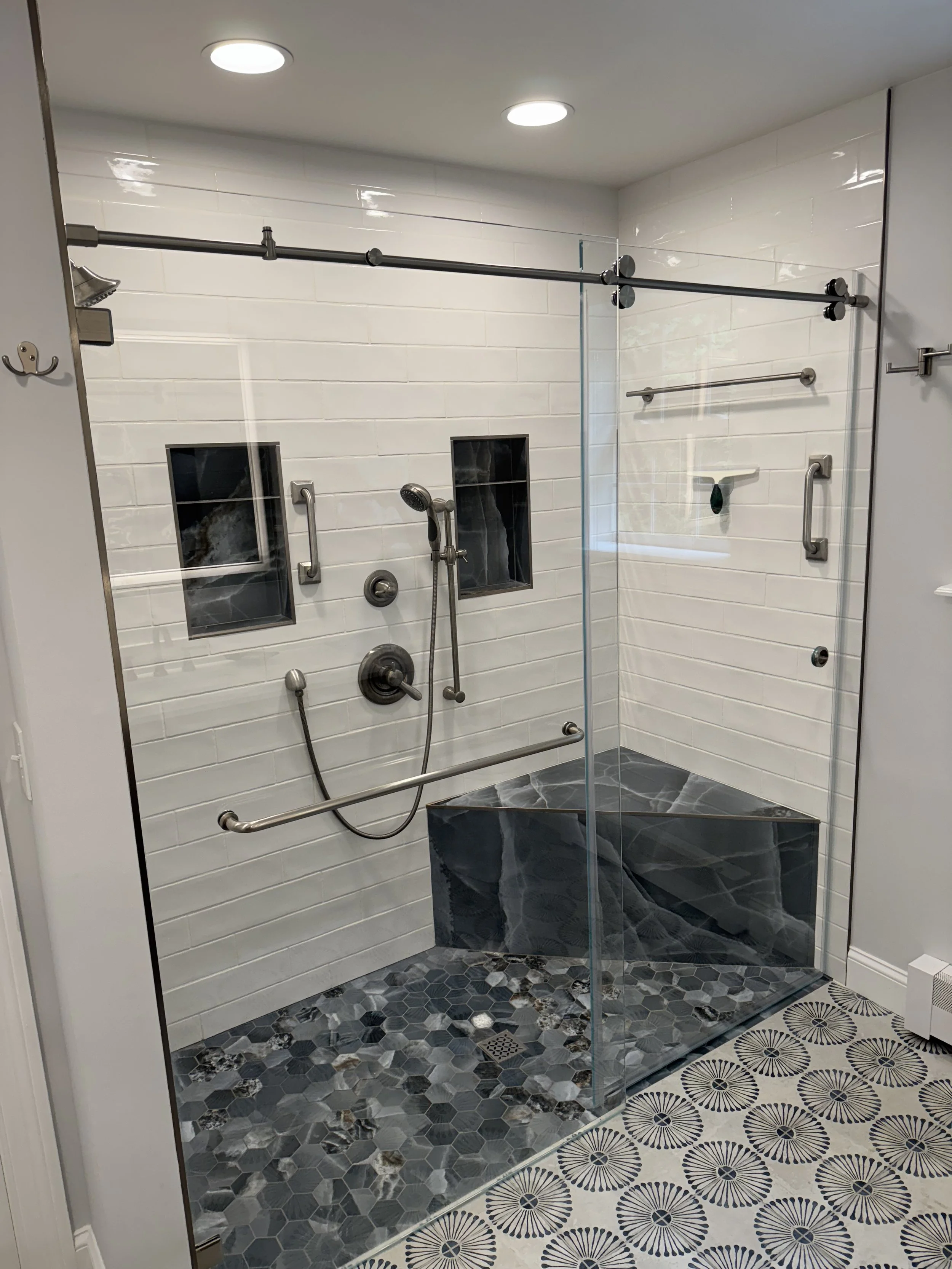 Bathroom shower with white subway tile walls, two small inset shelves, a handheld shower head, and a bench with dark veined gray stone. The shower has a glass door and adjacent patterned tile floor.