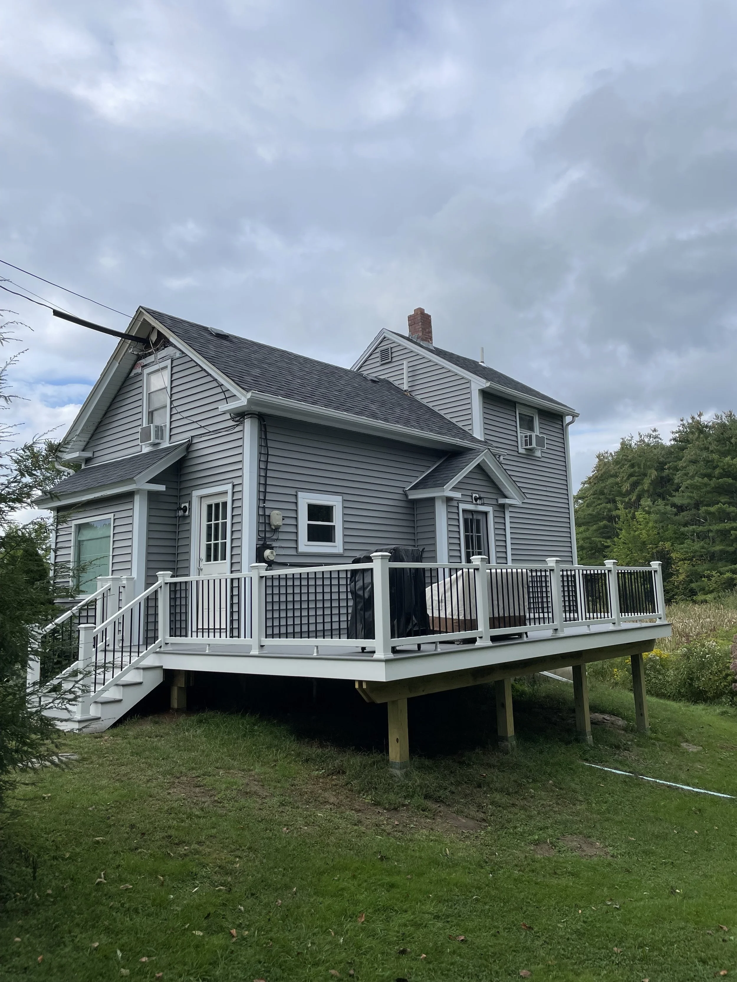 A two-story gray house with white trim and a deck in a yard with green grass and trees, under a cloudy sky.