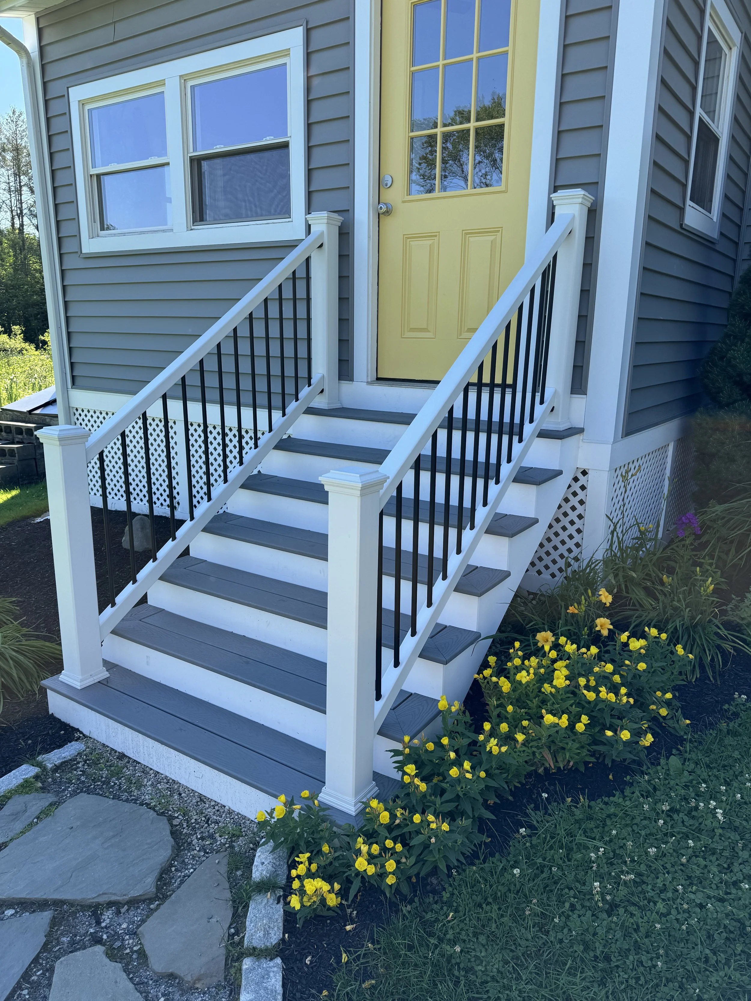 Front porch stairs leading to a yellow door on a blue house, with flower beds and yellow flowers in the garden.