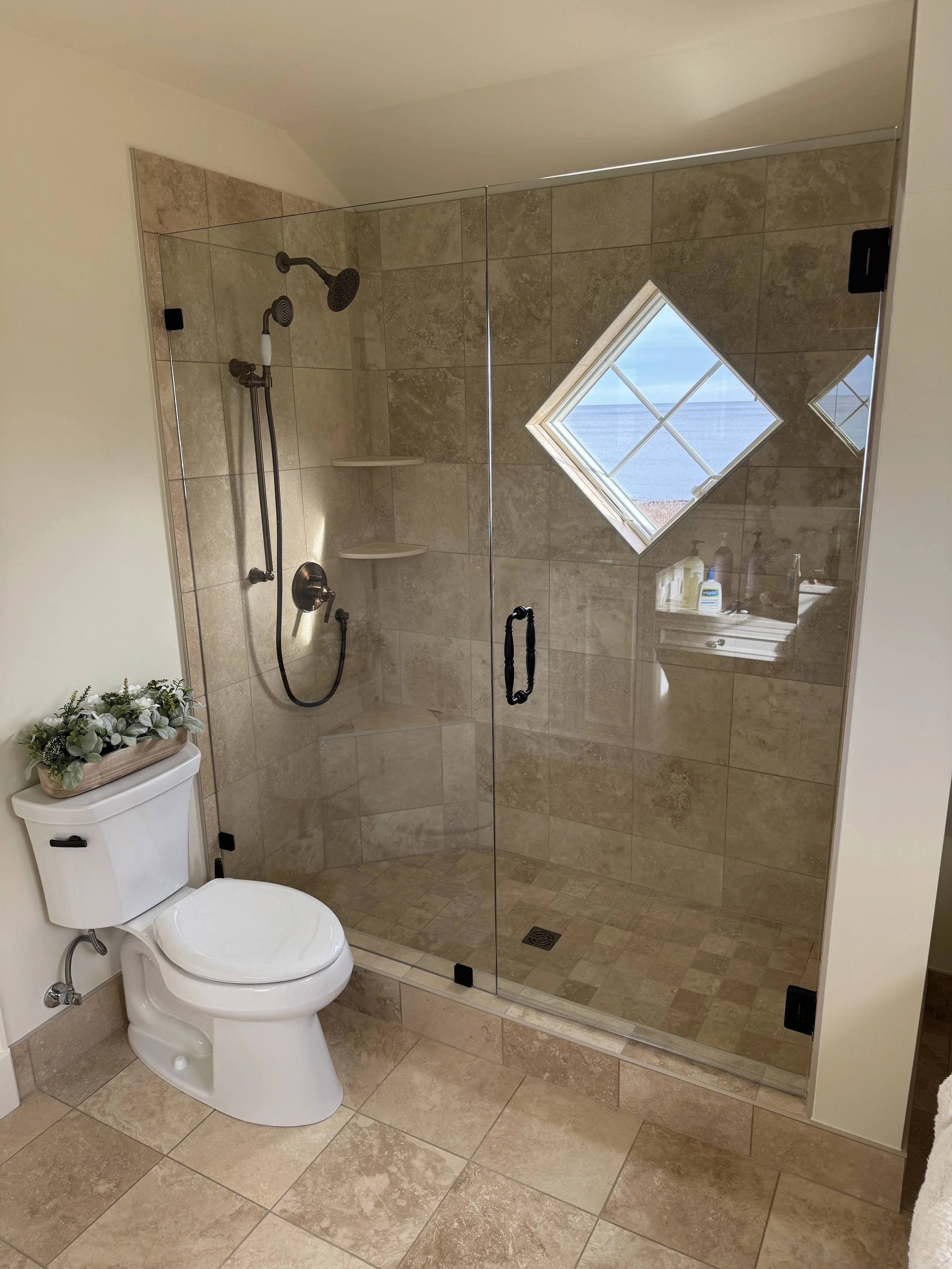 A bathroom with glass shower enclosure, beige tiled walls and floor, a small window showing an ocean view, and a white toilet with a decorative plant on top