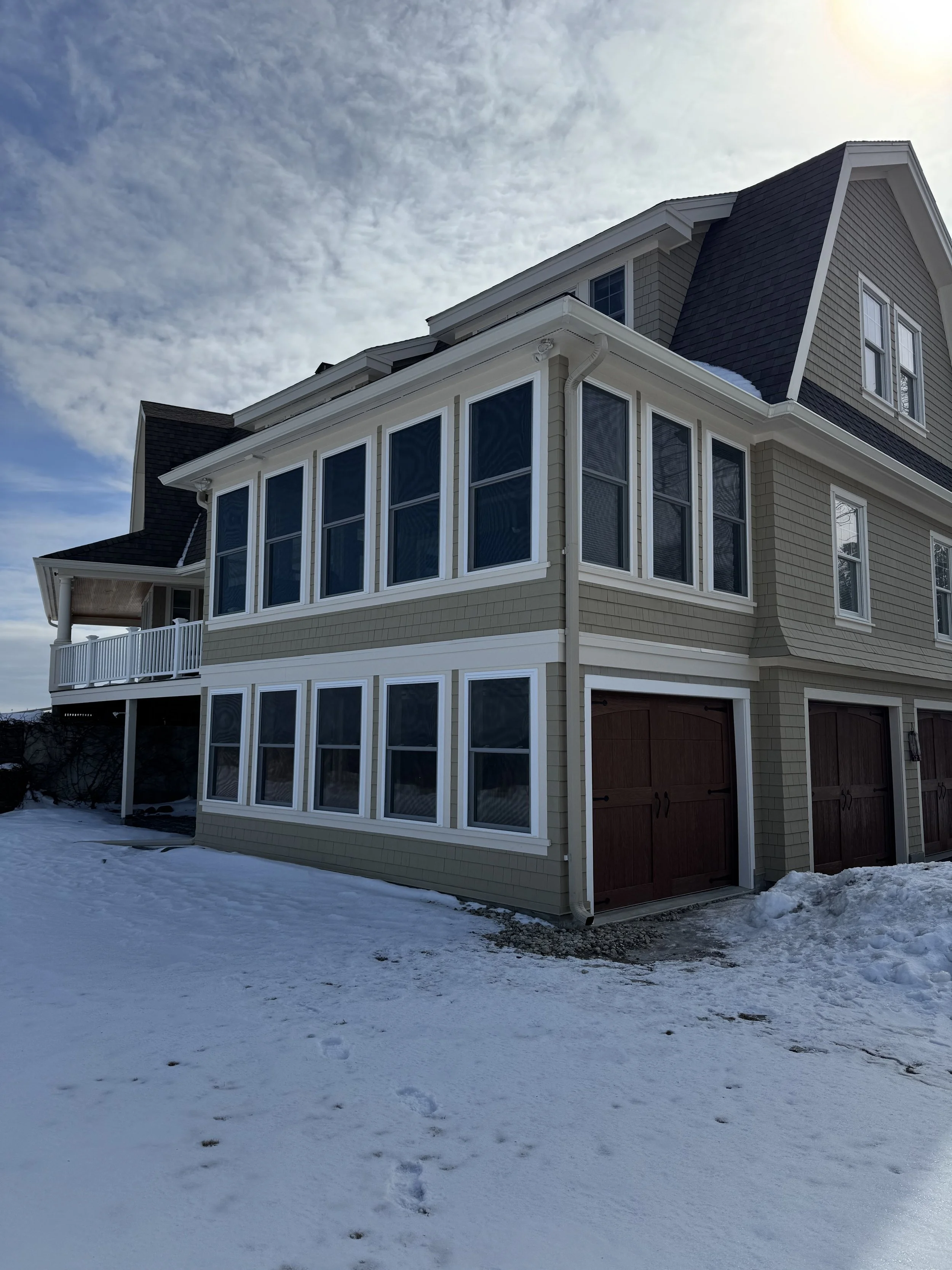 A multi-story house with beige siding, white trim, and a dark roof, surrounded by snow on the ground and under a partly cloudy sky.