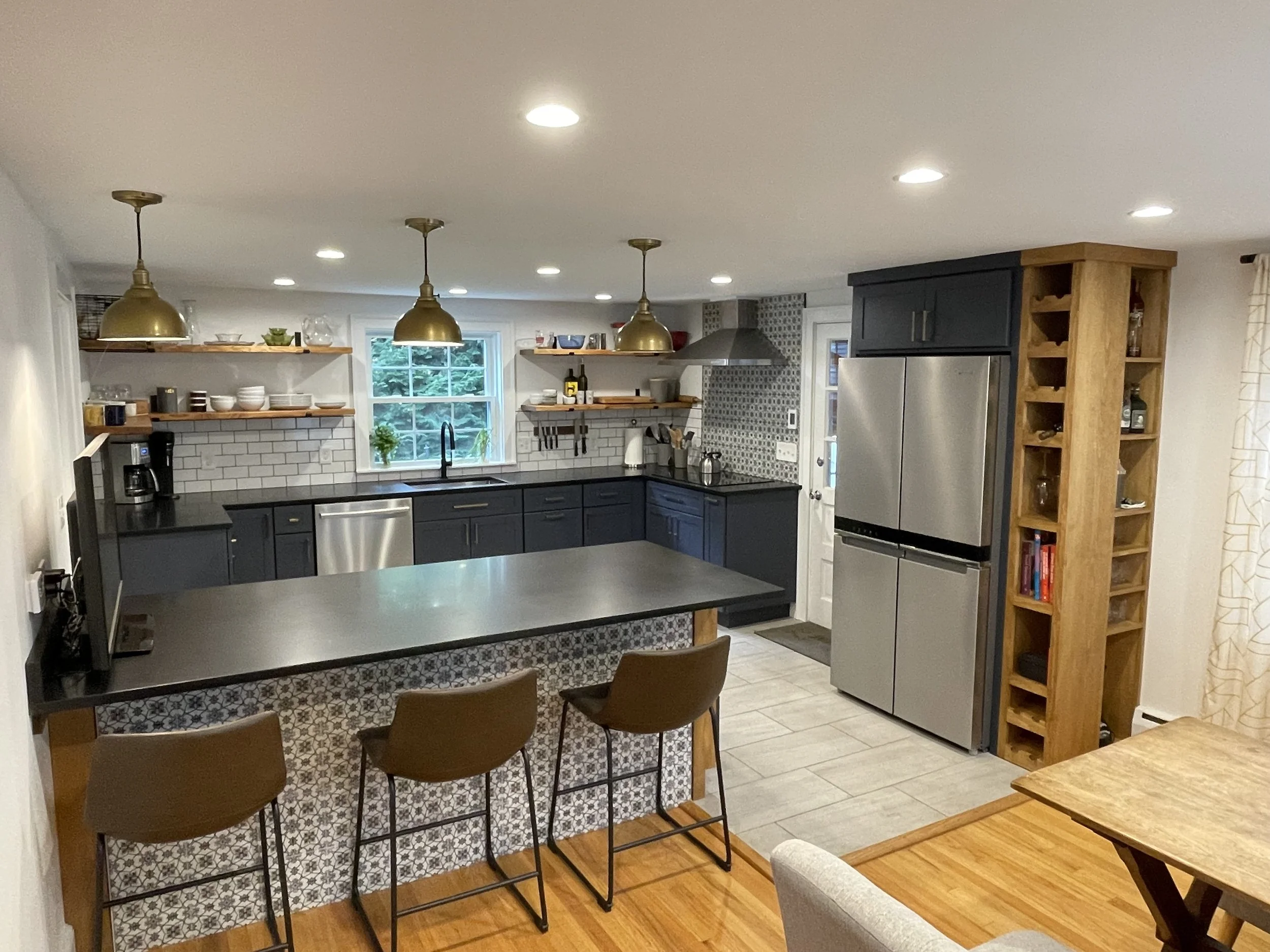 Modern kitchen with black cabinets, stainless steel appliances, open shelving with dishes, and a breakfast bar with three brown chairs.