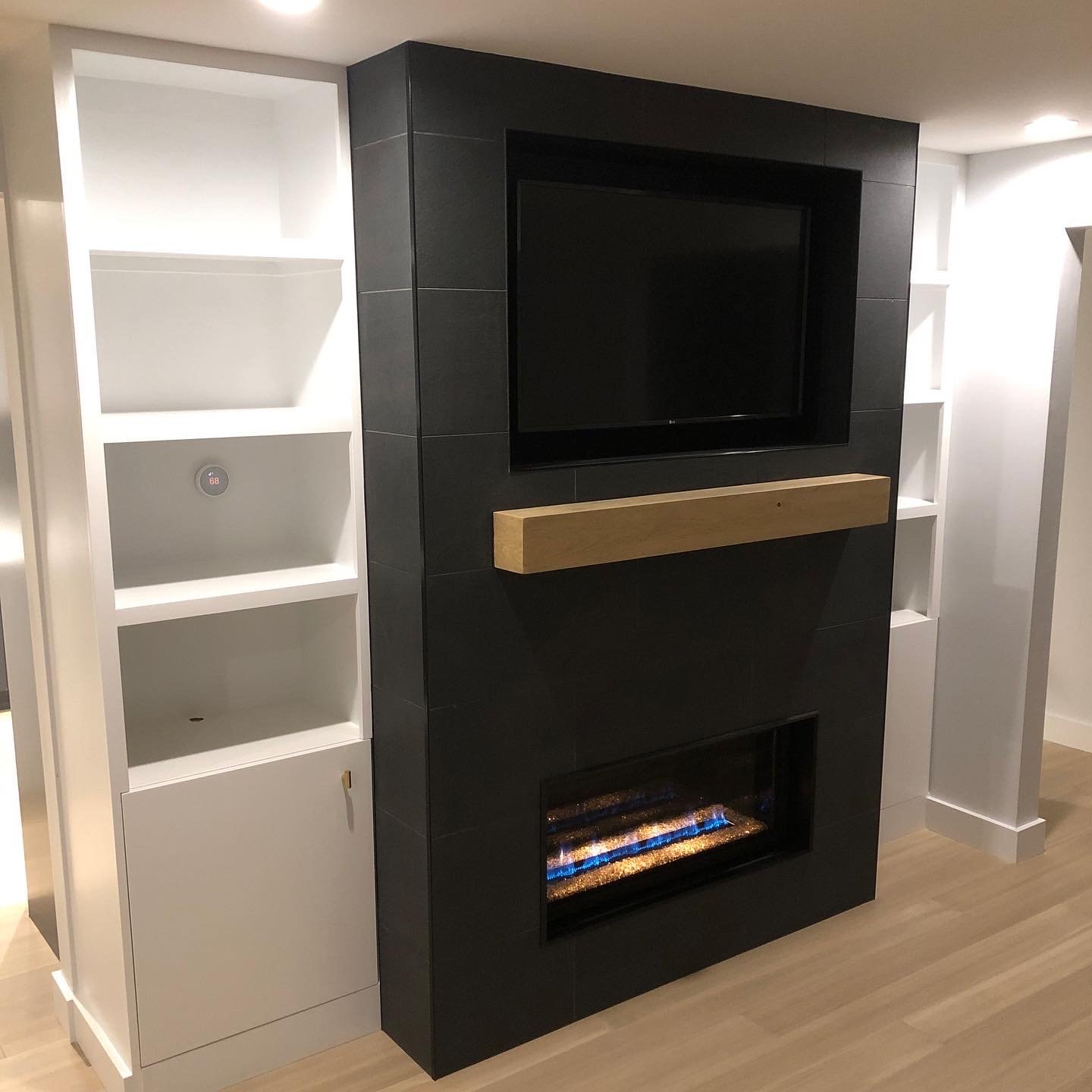 Living room corner with a black entertainment wall, mounted flat-screen TV, electric fireplace, wooden trim, and white built-in shelves with a digital thermostat.