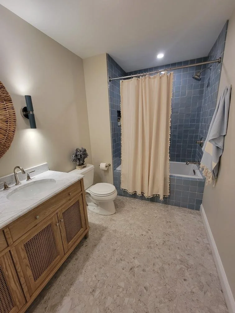 A bathroom with a wooden vanity, marble countertop, and a mirror, a toilet, and a shower with blue tiles and beige curtain.