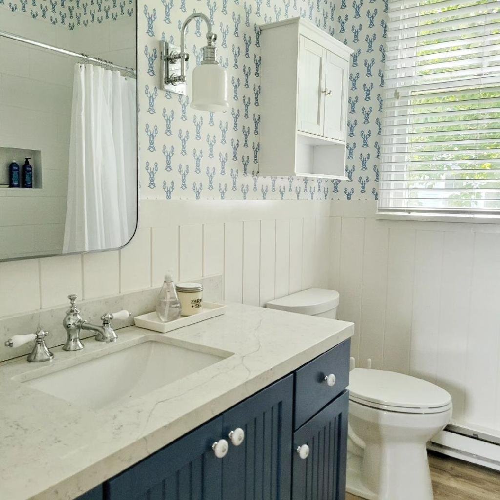 A bathroom with a white marble countertop, a navy blue vanity, white paneled walls, and a wallpaper with blue cactus patterns. There is a mirror, a wall-mounted light fixture, a cabinet above the toilet, a window with white blinds, and a visible shower area with a white curtain.
