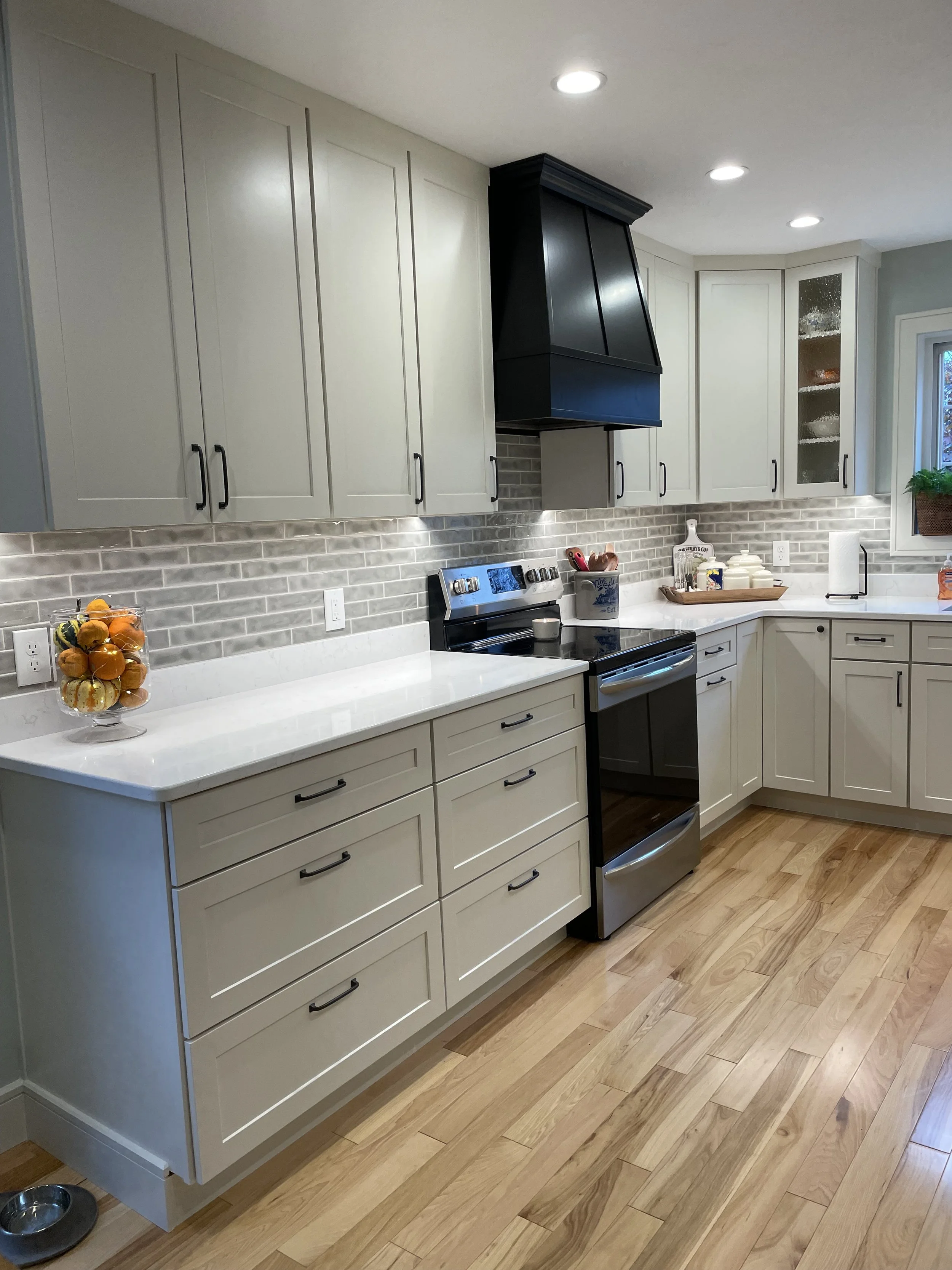 Kitchen with white cabinetry, wooden flooring, gray brick backsplash, black range hood, stainless steel oven, and fresh lemons in a glass bowl.