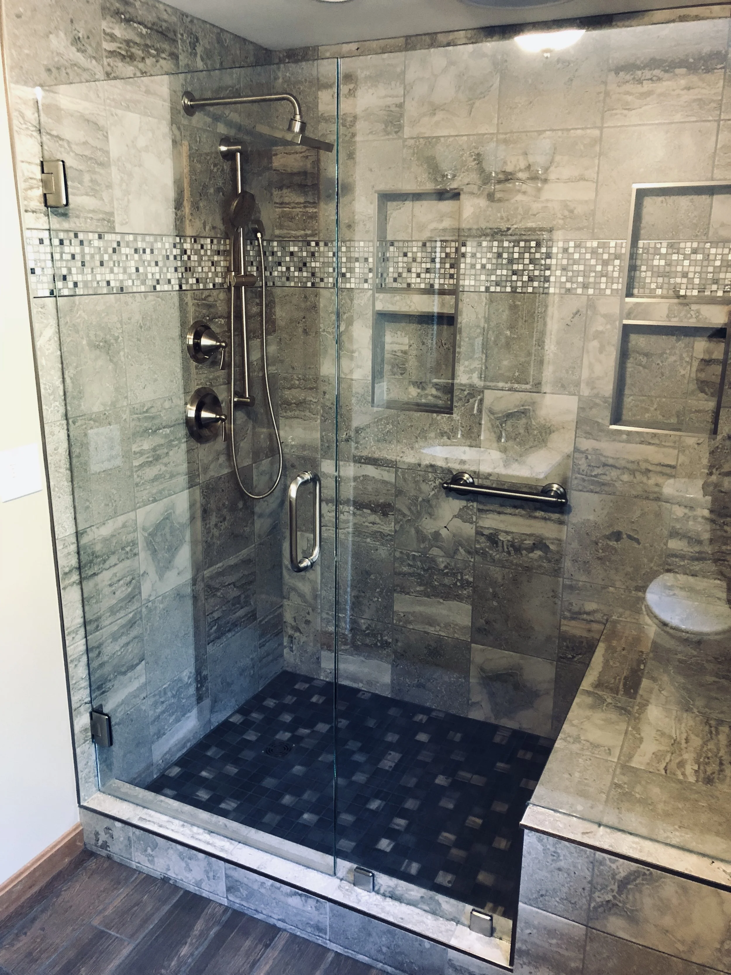 A modern glass-enclosed shower with gray and beige stone tiles, a mosaic border, built-in shelves, and a black and gray tiled shower floor.