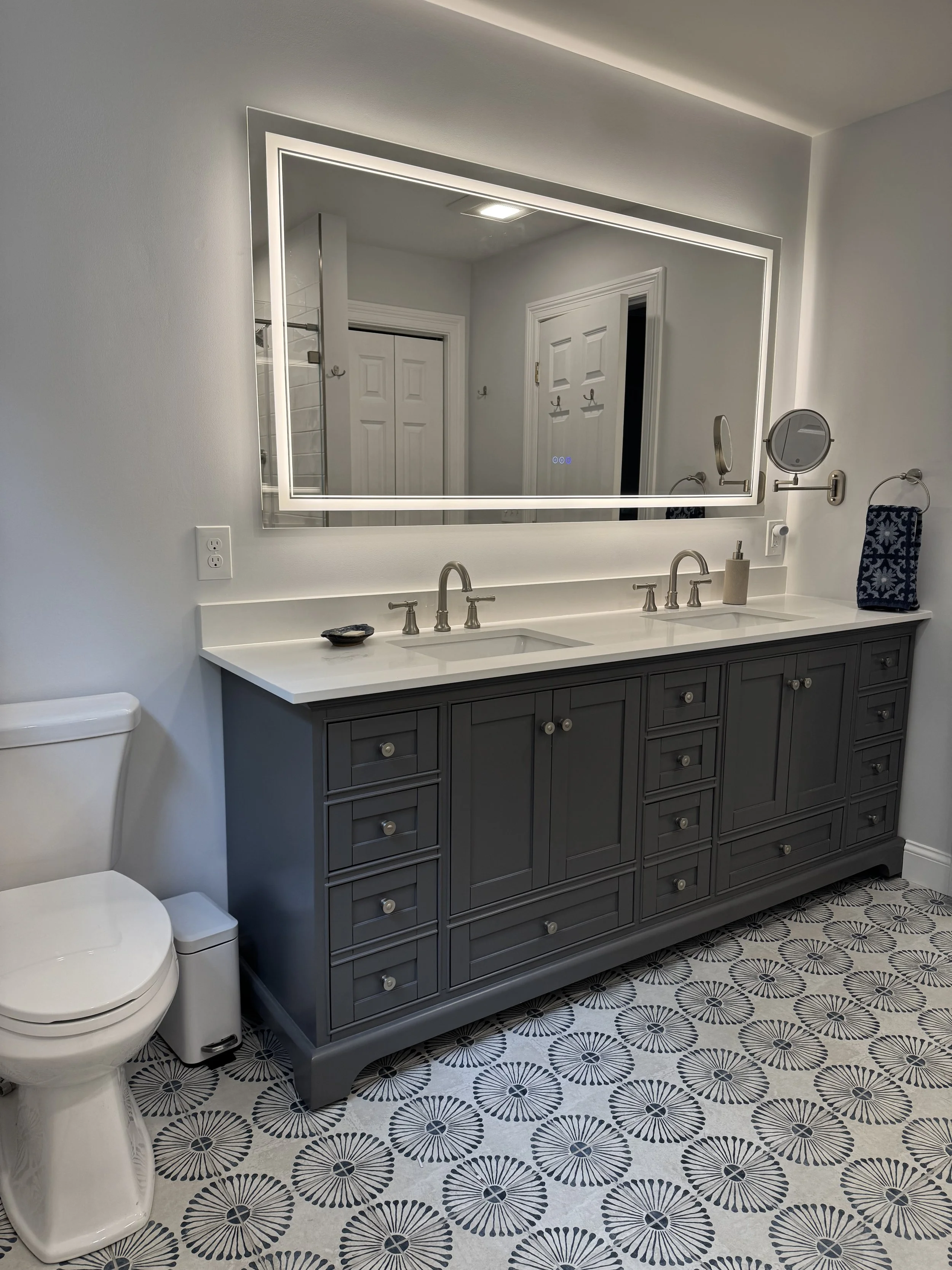 Modern bathroom with white walls, a large illuminated mirror, a double vanity with a gray cabinet and white countertop, and patterned floor tiles.