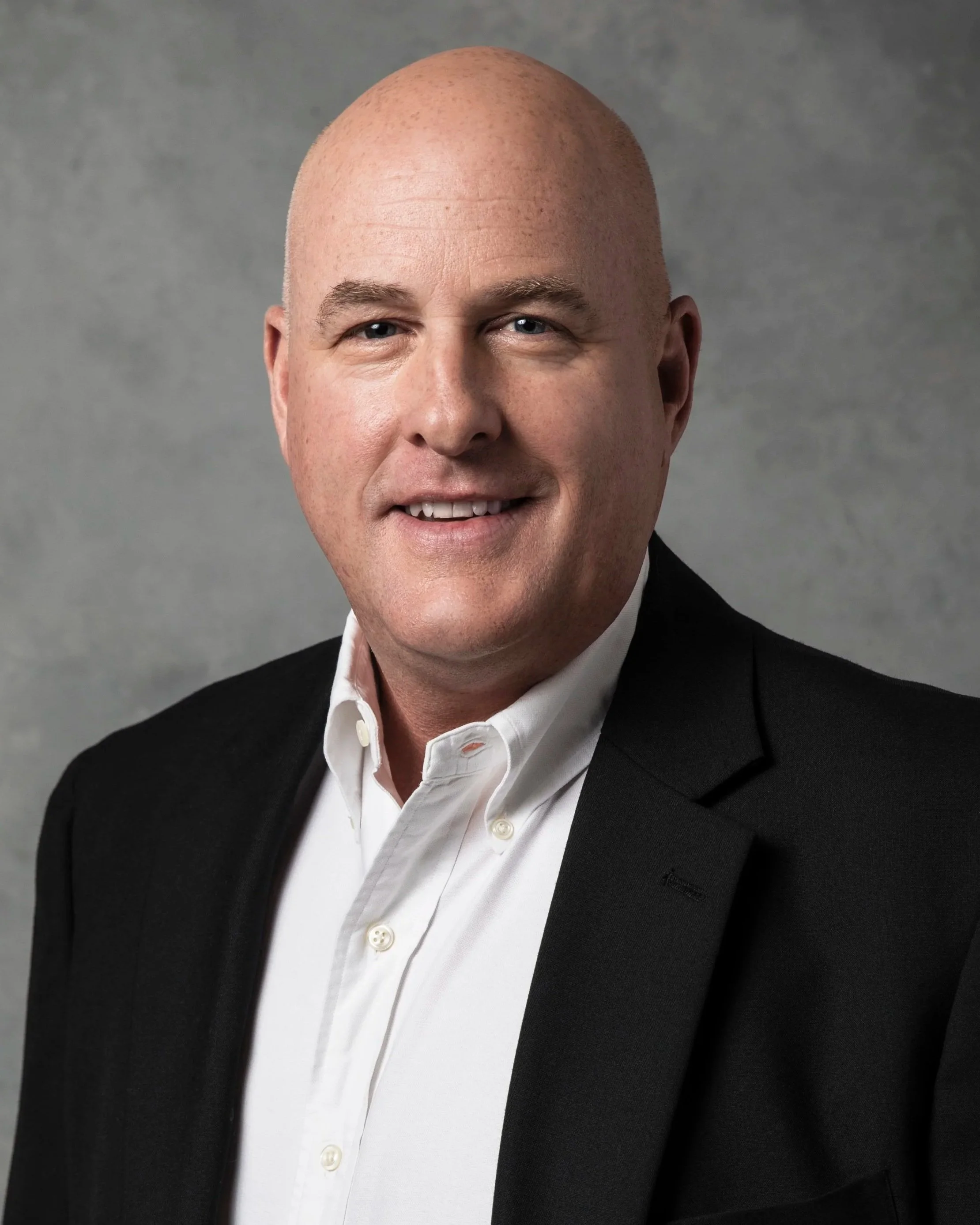 A professional headshot of a bald man in a black suit and white dress shirt, smiling against a grey background.