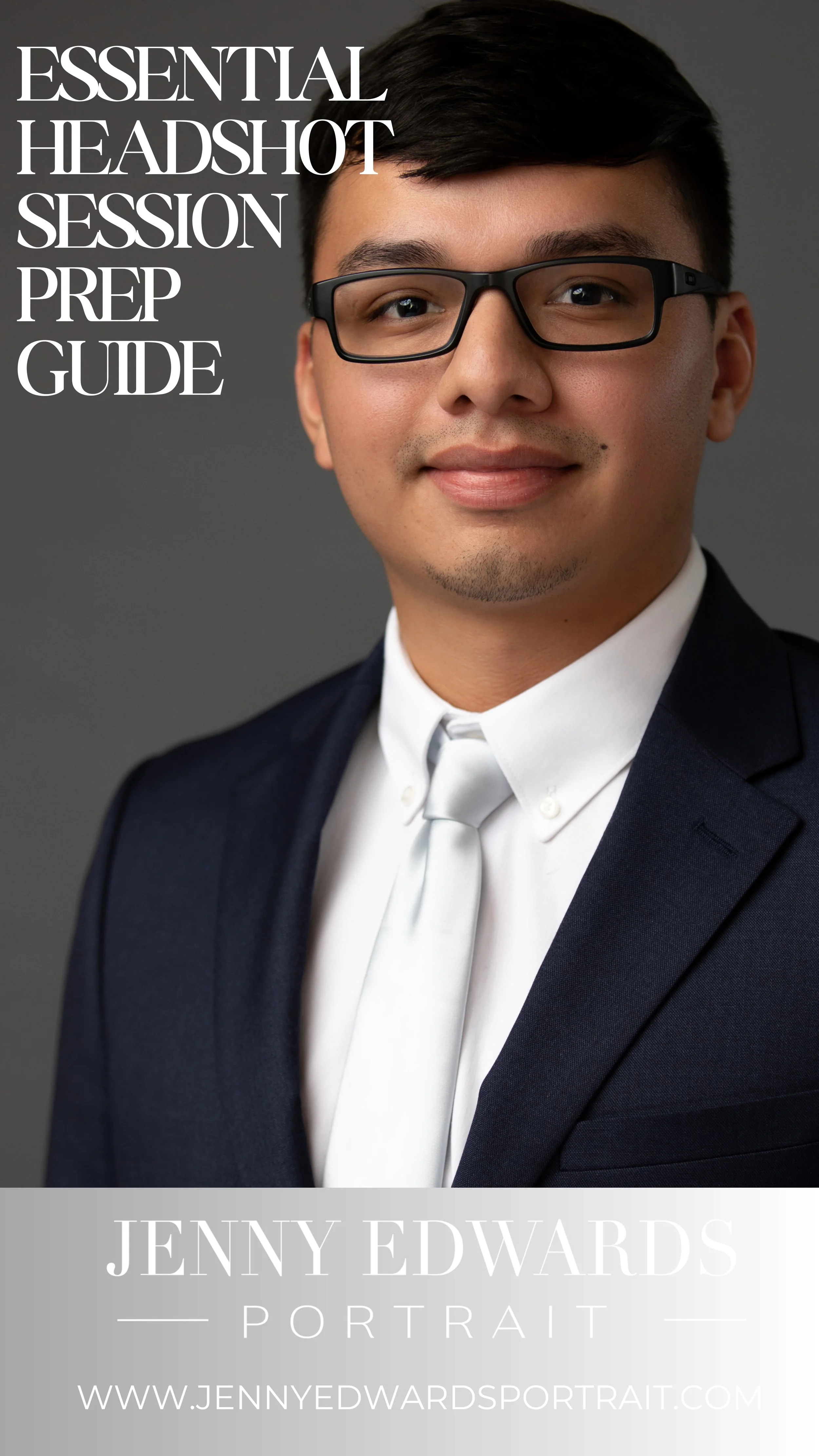 A professional headshot of a young man with glasses, wearing a dark suit and white shirt with a tie, smiling in front of a gray background.